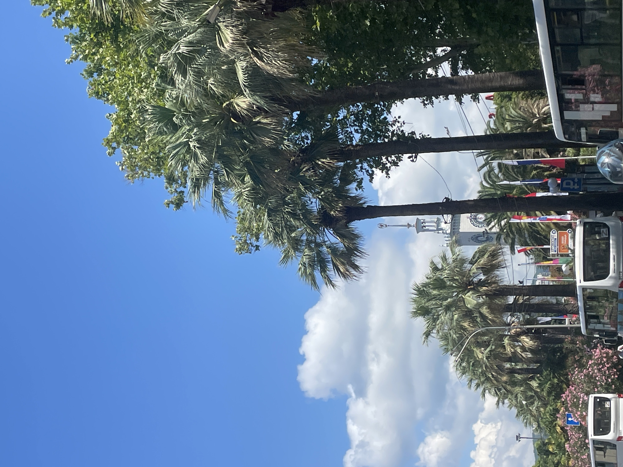 A low-angle view captures a street scene in Sochi, Russia, with a clear blue sky dotted with fluffy white clouds. A line of tall palm trees dominates the right side of the frame, their fronds casting shadows on the scene below. In the background, a distinct white clock tower with a spire is visible, a landmark that suggests the location is near the Sochi Sea Port. Flags of various nations are displayed along the street, fluttering in the breeze. Buses are present on the road, indicating active transportation within the city. The scene is bathed in bright sunlight, suggesting it was taken during the daytime.