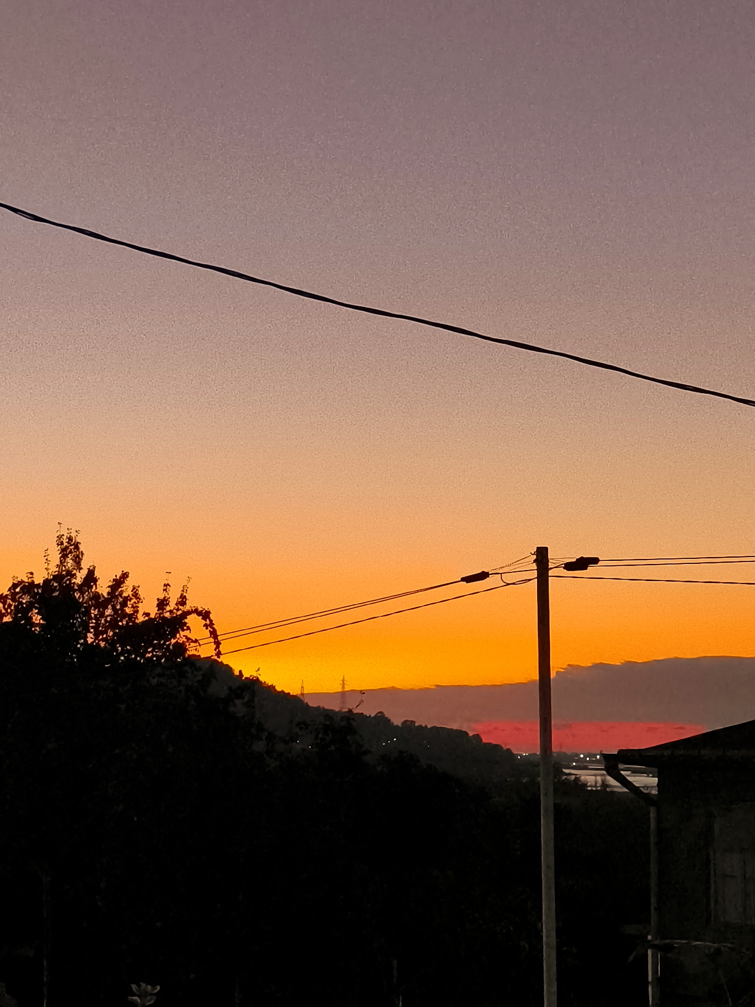 The image depicts a sunset over a landscape that includes elements of a town or city. The sky transitions from a pale purple at the top to a vibrant orange and red towards the horizon, indicative of a sunset. A dark silhouette of trees and hills occupies the lower portion of the image, suggesting a natural or semi-natural setting. Man-made structures are also visible, including utility poles with wires, and what appears to be part of a building on the right side. In the distance, faint lights suggest the presence of a settlement or urban area. The overall impression is one of a dusk scene in a location that blends natural elements with human infrastructure. It is not possible to confirm the location as Makho, Georgia, based solely on the visual information provided in the image.