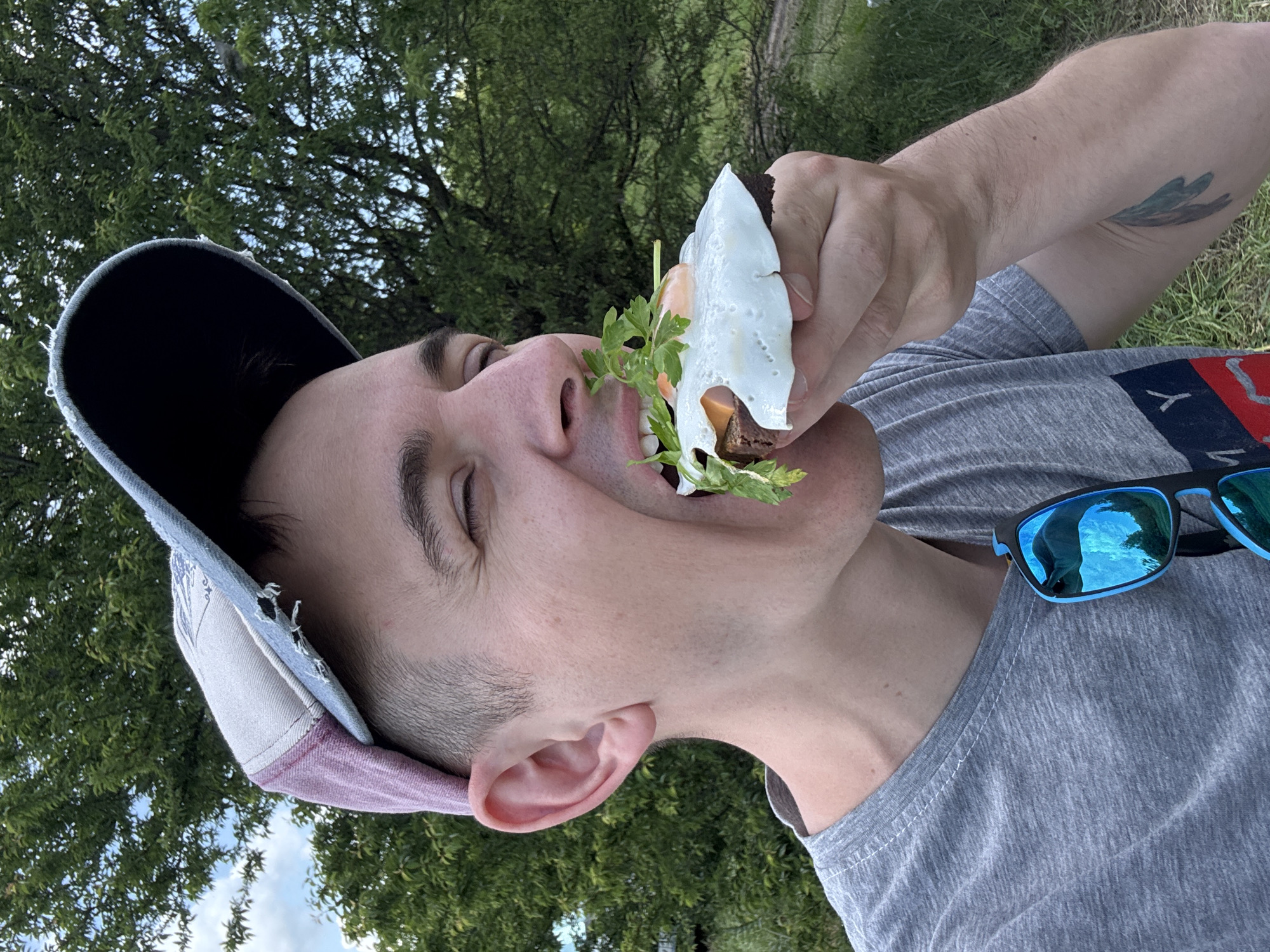 A man wearing a grey t-shirt and a baseball cap with a dark brim is holding a sandwich up to his mouth. He is outside in a grassy area with trees visible in the background. A pair of blue sunglasses is hooked onto the collar of his shirt. The location cannot be determined from the image.