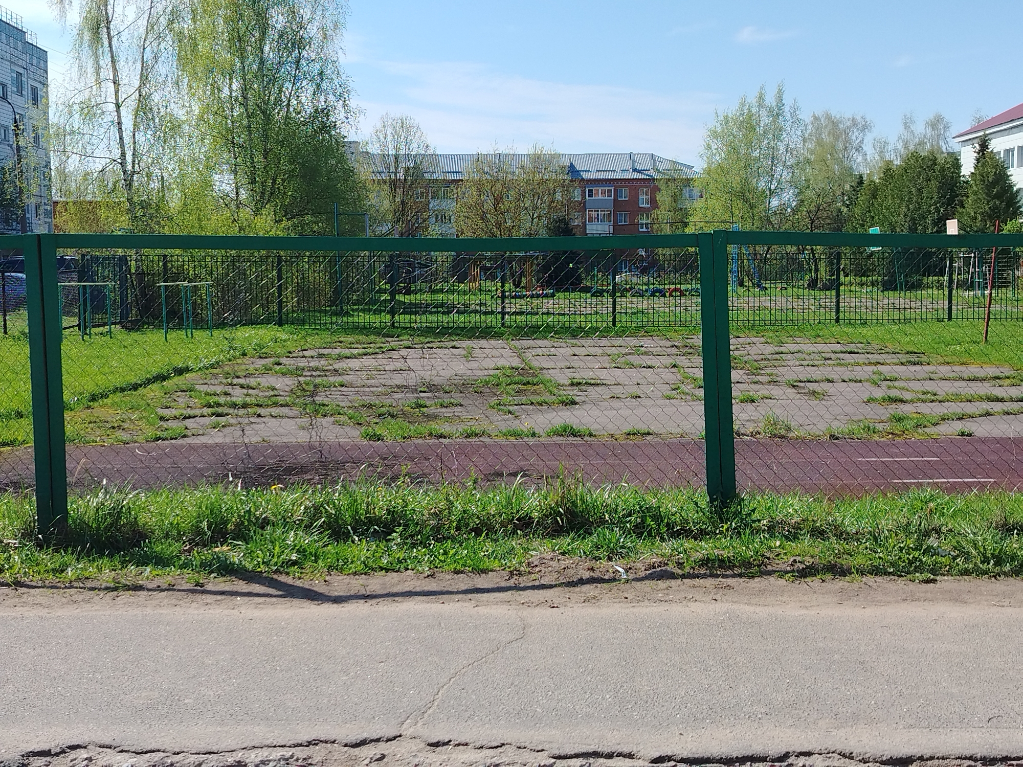 The image shows a fenced-off area with damaged pavement. Patches of grass grow through cracks in the pavement. There's a green metal fence in the foreground. Beyond the fence, there's a playground with various equipment and patches of grass. In the background, buildings are visible with trees surrounding them. A street runs along the front of the image. The location is Unknown, Unknown. The sky is blue with few clouds.