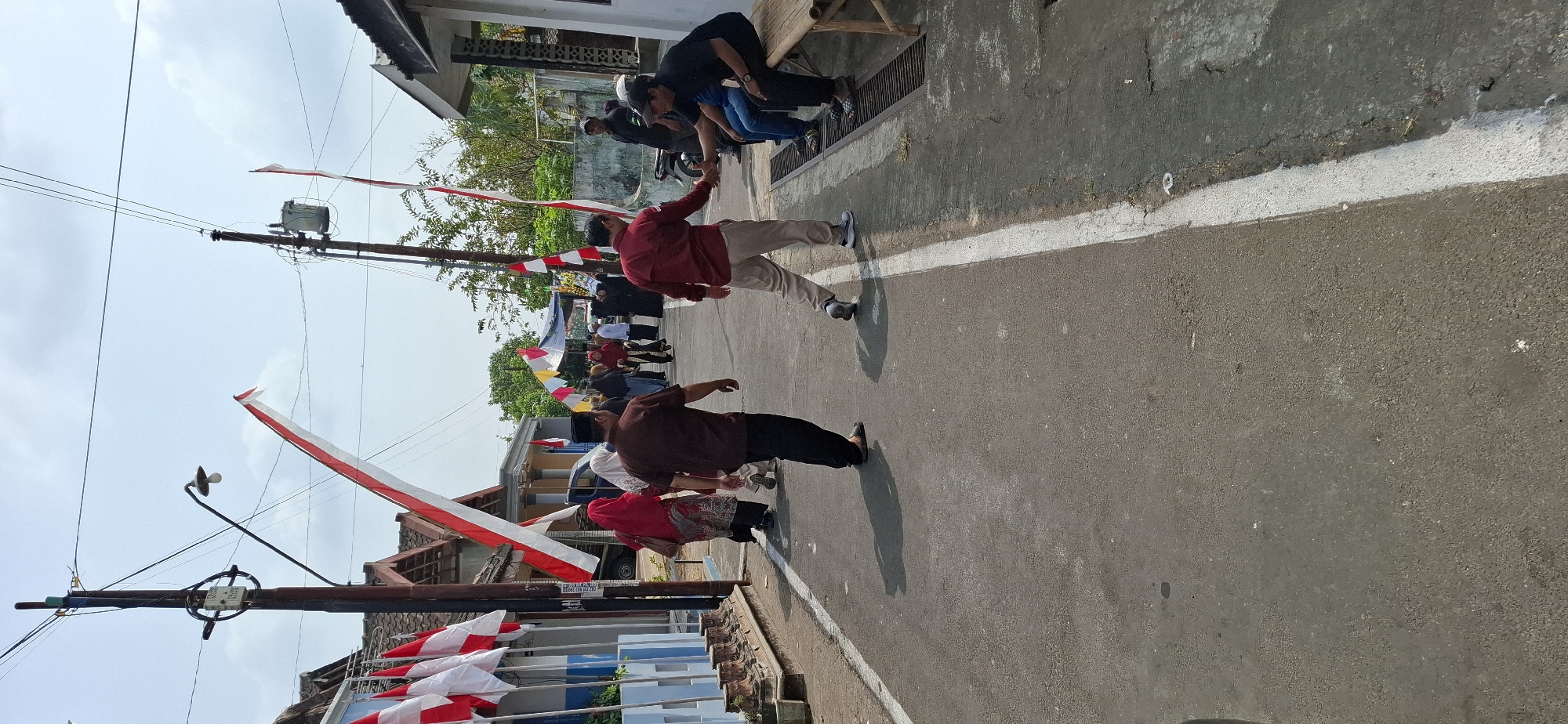 In the city of Gentan, Indonesia, on a sunny day, a street scene unfolds with residents and visitors participating in a local event. The road, paved with asphalt and marked with a white line, is lined with houses and adorned with Indonesian flags. A group of people are seen walking along the street, some wearing traditional attire, while others are casually dressed. In the foreground, two men are engaged in a handshake, possibly a greeting or a formal acknowledgement. To the right, a few individuals are seated on a wooden bench, observing the surroundings. The presence of electrical poles with overhead wires indicates the urban or semi-urban setting. The atmosphere appears festive, with flags adding a touch of national pride to the occasion.