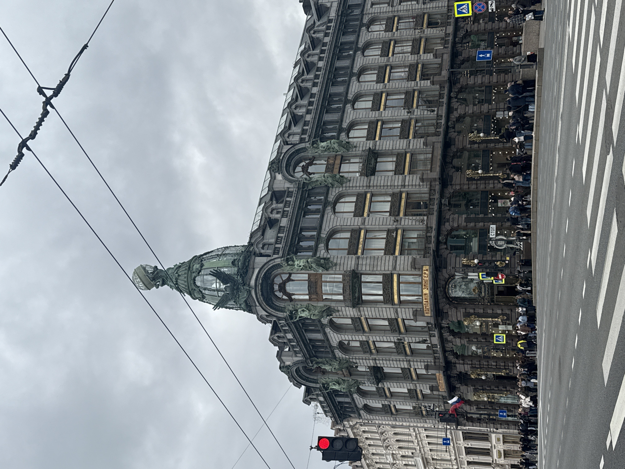 The image captures the Singer House in Saint Petersburg, Russia, as seen from street level. The architectural facade is detailed with sculptures and ornate window frames. A dome with sculptures is prominently featured on the building's corner. Power lines are visible against the overcast sky. A pedestrian crosswalk extends into the foreground. People are present near the building's entrance. A traffic light is red.