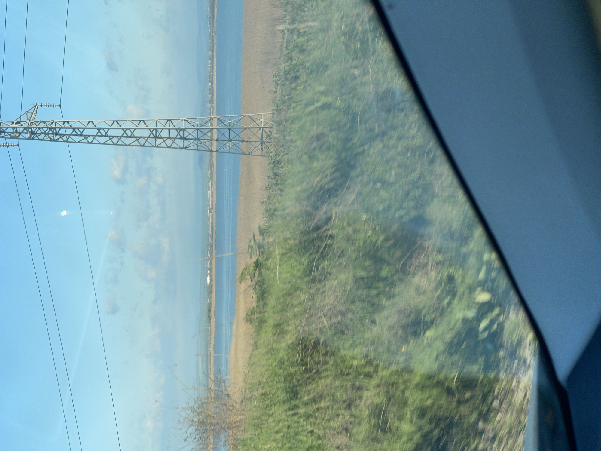 The image appears to be taken from inside a moving vehicle, possibly a train, looking out of a window. A large metal electricity pylon is visible on the left of the frame, with power lines stretching across the clear blue sky. The pylon is positioned between the vehicle's window and the sea. Beyond the pylon, there is a body of water, possibly the Black Sea, bordered by a narrow strip of land. The shoreline appears to have some buildings along it. The foreground shows overgrown vegetation.
The scene could be located in the vicinity of Burgas, Bulgaria, since that area is known for its coastal location and the presence of industrial infrastructure, including power lines and pylons running along the coastline. However, it's impossible to confirm the exact location based on this image alone.