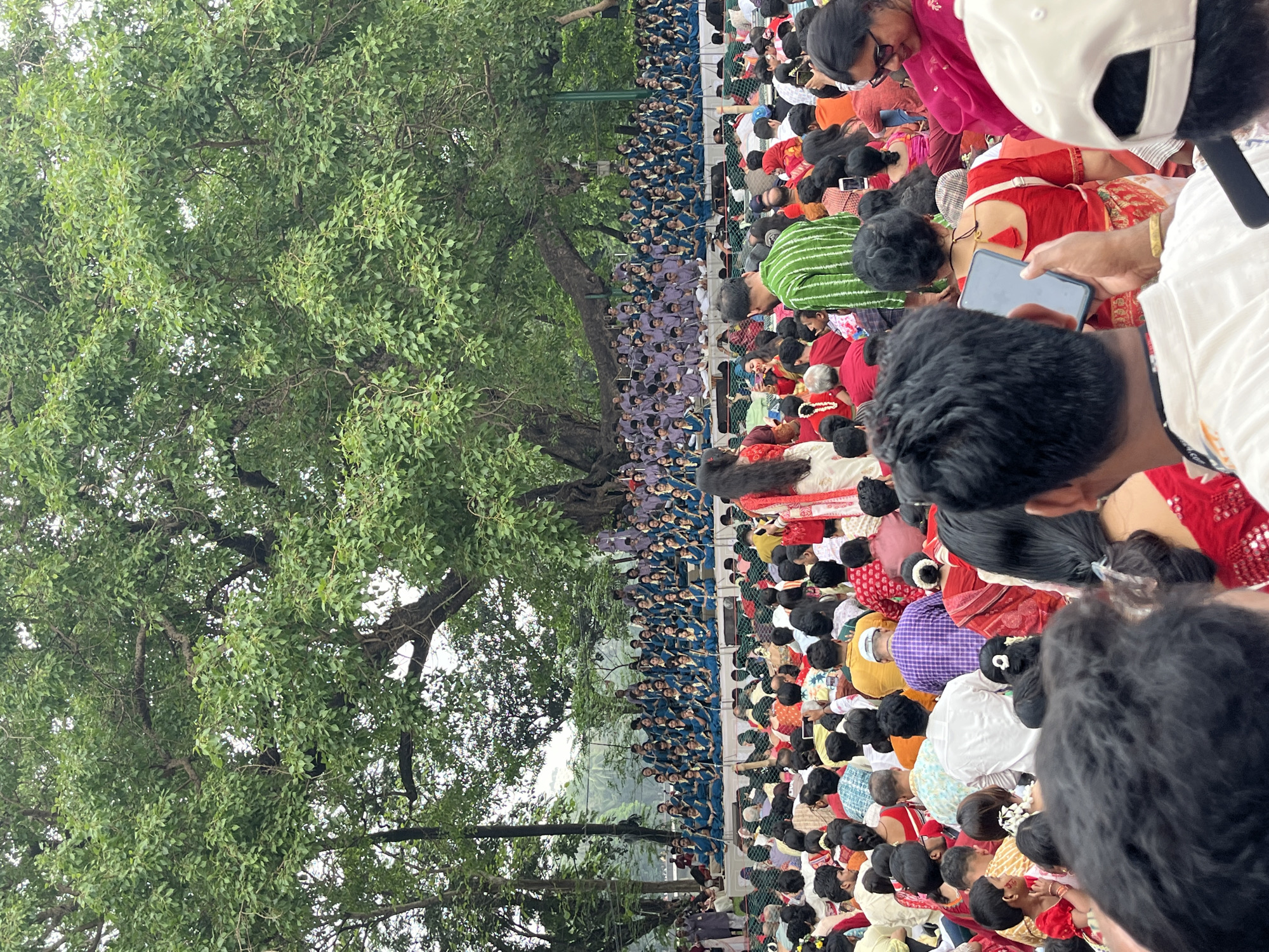 A large group of people are gathered outdoors, possibly in a park or open space. A dense tree with green leaves occupies a significant portion of the left side of the image. A large group of people wearing blue uniforms are gathered. Other people are in the foreground, wearing various colorful clothing, sitting or kneeling. There is a person taking a photo of the event. This could be a public event, performance, or gathering. This takes place in Dhaka, Bangladesh.