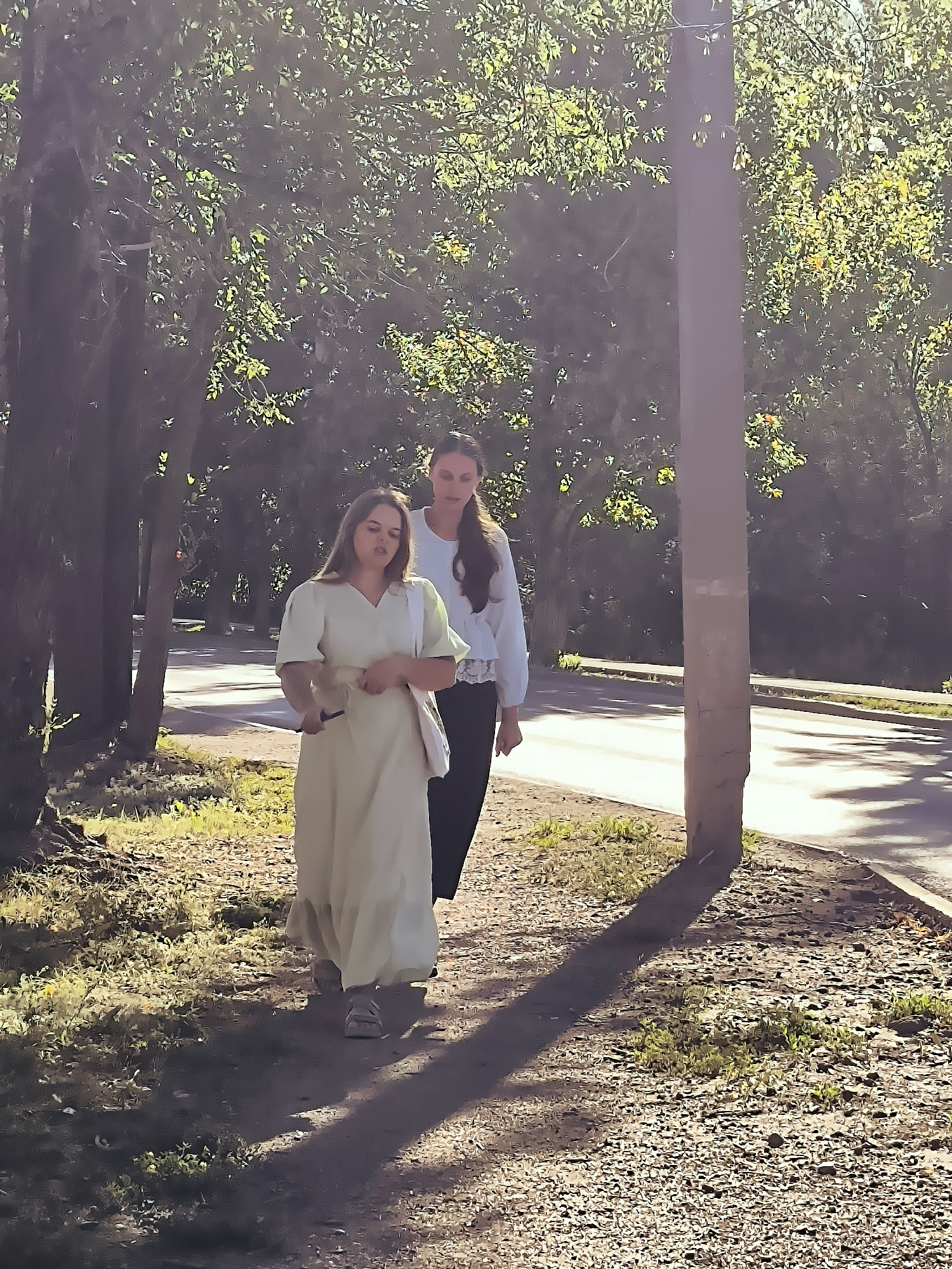 Two women are walking on a gravel path next to a paved road. The woman in the foreground is wearing a long, light-colored dress and sandals, and she is holding a phone in her left hand and a white bag over her right shoulder. The woman behind her is wearing a white blouse with ruffled details and dark pants. They are walking in the same direction, and the sun is casting shadows. The environment appears to be a park or a tree-lined street, with trees on the left and foliage on the right. A utility pole is visible on the right side of the image. The scene is set in the city of Qarağandı, Kazakhstan, though no specific landmarks of the city administration are clearly identifiable in this image.