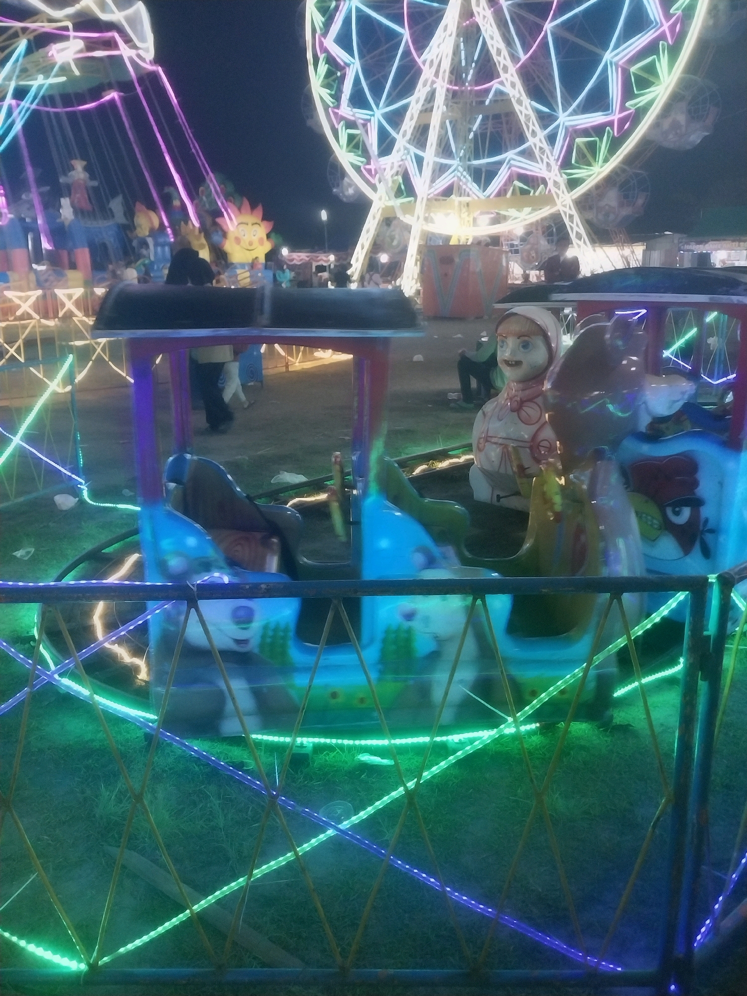 In Jiwut, Indonesia, at night, an amusement park is illuminated by colorful lights. In the foreground, a small amusement ride with multiple carriages is visible. One carriage features a doll-like figure with a white head covering and blue eyes, wearing a red and white outfit. Other carriages appear to be decorated with cartoonish animal faces, including a panda and a bird. The ride is surrounded by a fence adorned with green and purple LED strip lights. In the background, a large Ferris wheel with white, green, and pink lights dominates the scene, along with a swing ride also lit with pink lights. Several people are visible in the background, some walking and others possibly operating or enjoying the rides. The ground is covered with grass.