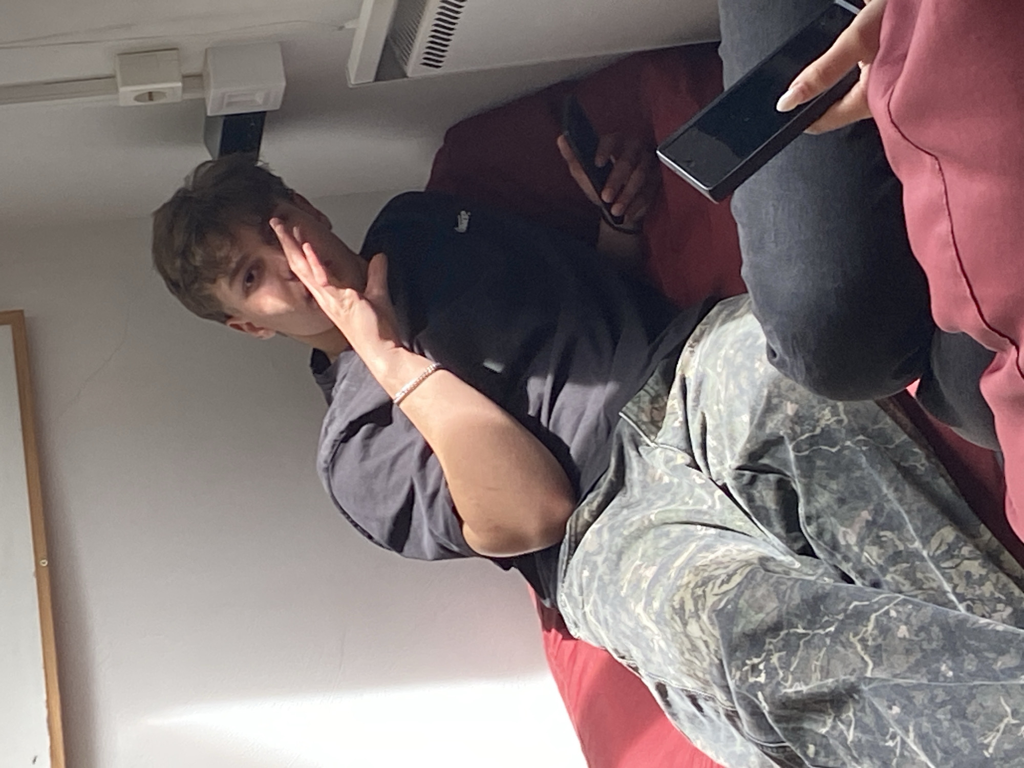 The image shows two people sitting on a red sofa. One person is covering their face with their hand, wearing a black t-shirt and patterned pants. A second person is sitting next to them, holding a phone, wearing a black shirt, jeans, and a red top. The background includes a white wall and ceiling with a radiator. The location is likely indoors. There is no clear indication of the city of Troyan, Bulgaria.