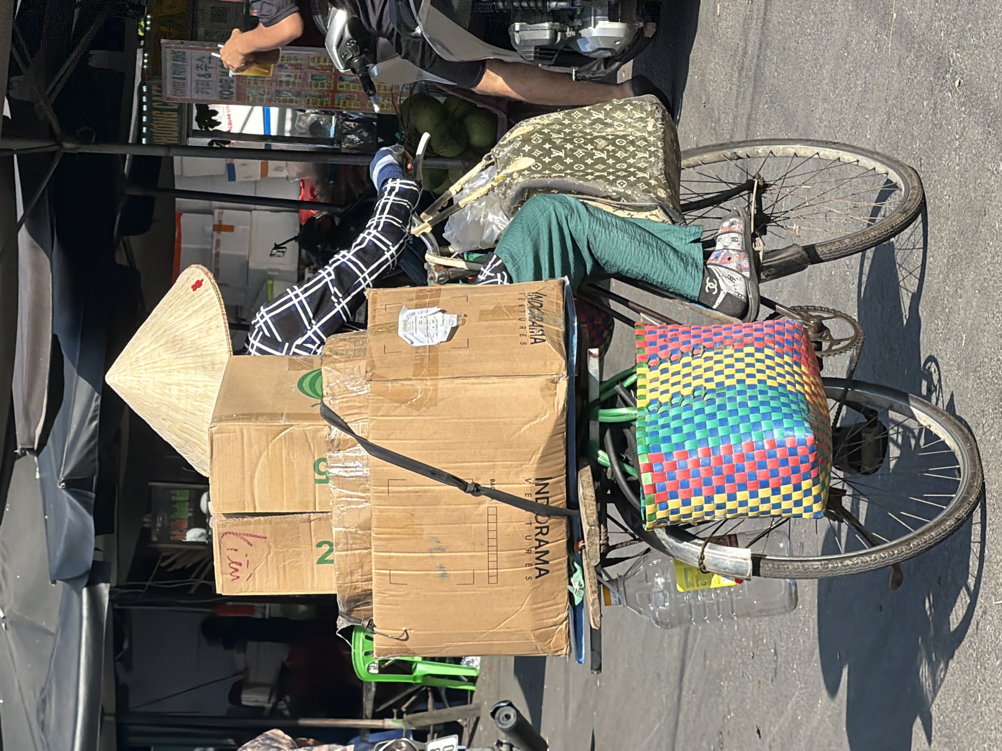 In Nha Trang, Vietnam, a person wearing a conical hat rides a bicycle loaded with several cardboard boxes. The boxes are stacked high on a flatbed attached to the rear of the bicycle. A large, patterned tote bag is slung over the back of the bicycle seat, and a brightly colored woven basket is attached to the rear wheel. A large plastic water bottle is strapped to the frame near the rear wheel. The person is wearing green pants and a long-sleeved shirt with a black and white checkered pattern. The bicycle appears to be in motion, with the person's right hand on the handlebar and their left arm resting on the boxes. The background shows a market setting with awnings and other stalls, suggesting a busy street scene.