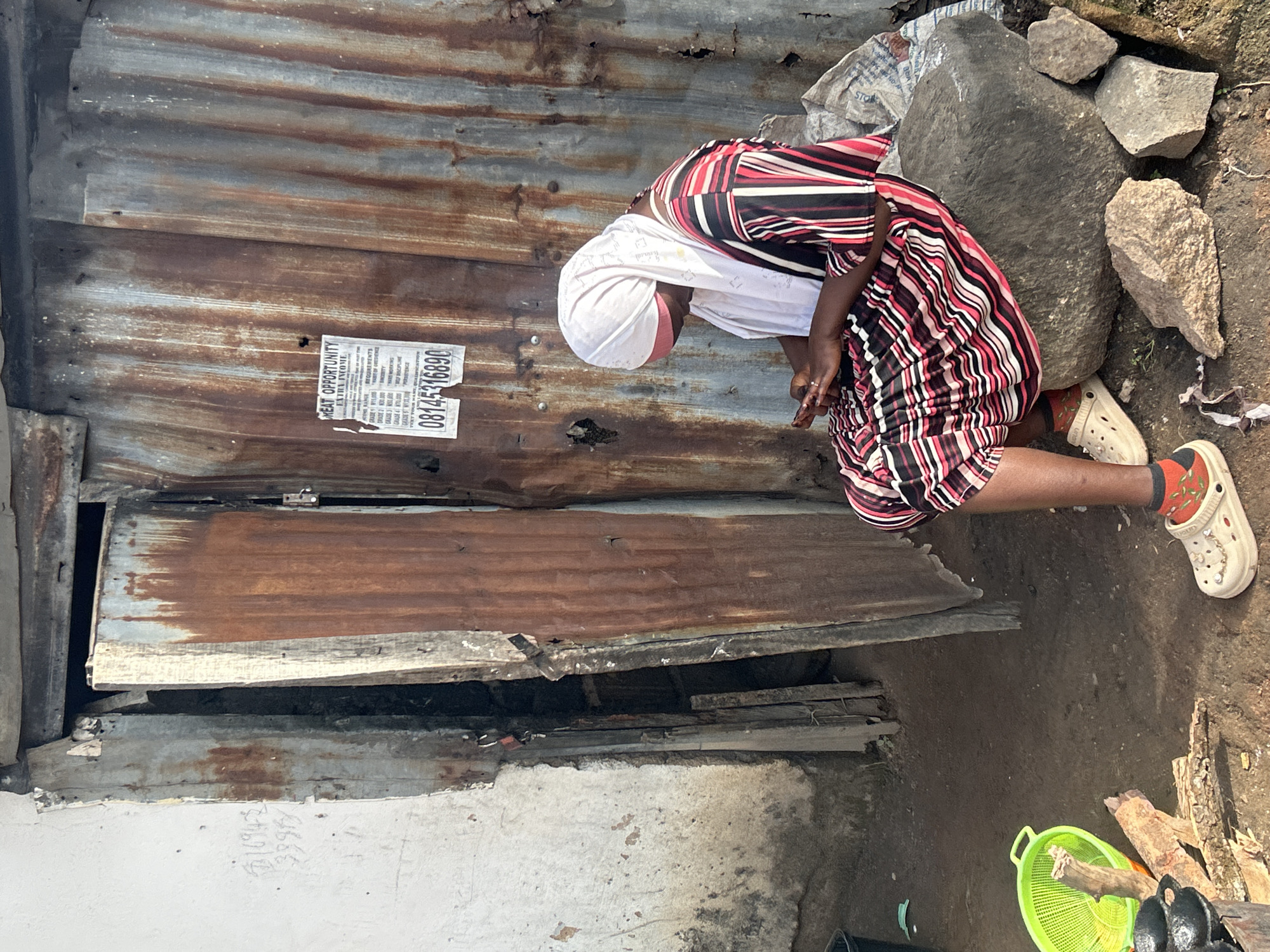 A woman sits on a large stone in front of a weathered corrugated metal structure, possibly a dwelling or shop, in Jos, Nigeria. She is wearing a white headscarf and a brightly striped dress. Her attention is focused on her mobile phone, which she holds in her hands. The background shows a rustic, somewhat dilapidated environment with an open doorway revealing shadows and debris. A poster with text and a phone number is affixed to the metal wall. In the foreground, a green colander, firewood, and cooking utensils are visible, suggesting domestic activity. The ground is dirt and scattered rocks.