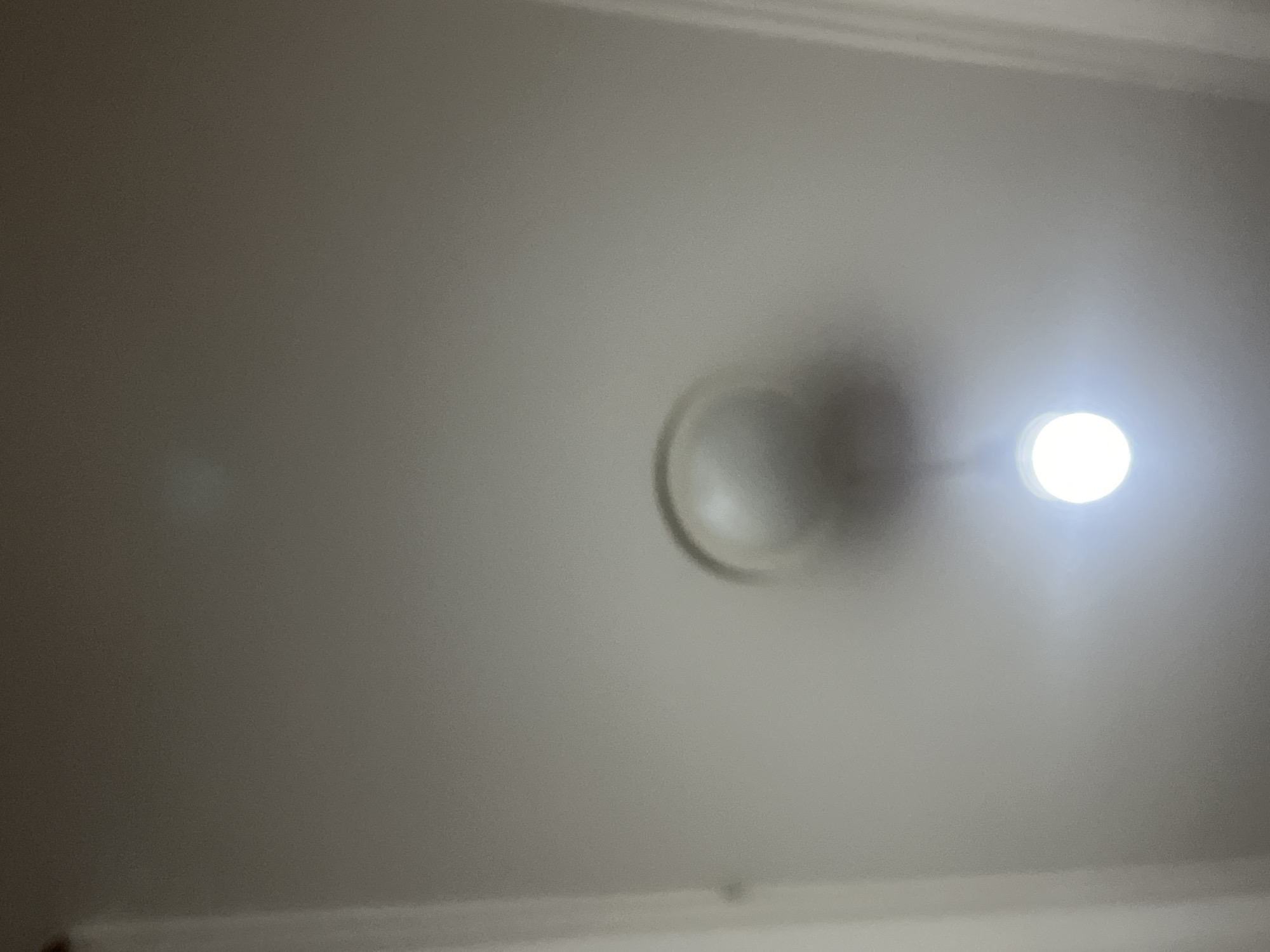The image is a low-angle, blurry shot of a ceiling light fixture. The fixture is a spherical globe with a visible lightbulb emitting a bright glow from within. The ceiling appears to be painted a neutral, light color. The image was taken in Jalingo, Nigeria.