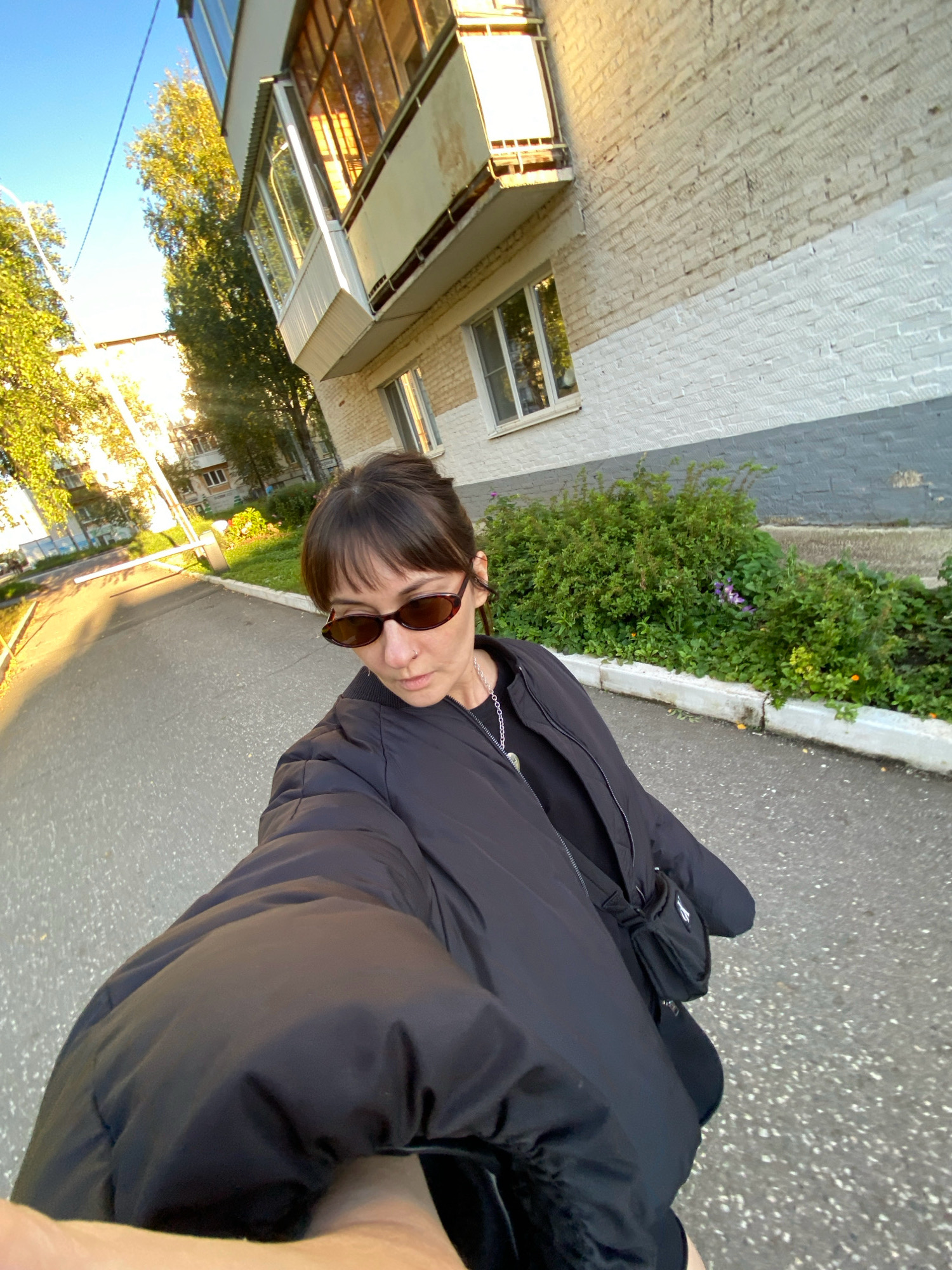 A young woman wearing sunglasses and a black puffer jacket takes a selfie outdoors on a paved street in Votkinsk, Russia. She has dark hair pulled back and is wearing a silver necklace. Behind her, a building with a balcony and brick facade stands against a clear blue sky. Trees and other buildings are visible in the background, and there is a small patch of green bushes with flowers in front of the building. The sunlight casts long shadows across the street.