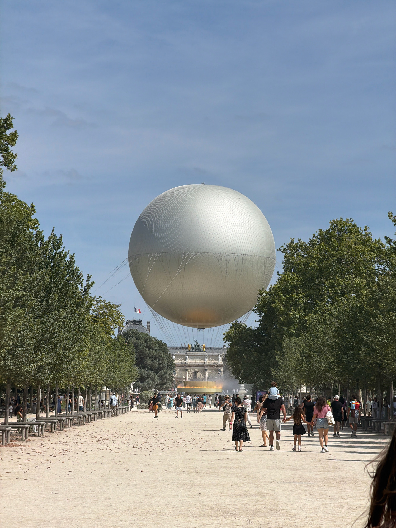 In the Tuileries Garden in Paris, France, a large silver balloon is tethered by cables and floats in a blue sky with scattered clouds. The balloon is positioned above a line of trees and a fountain. A crowd of people strolls along a wide, sandy path in the foreground, some walking in groups and others individually. The path is lined with trees on both sides, and benches are visible along the left side. In the background, a grand building with a dome and statues is visible, with the French flag flying from its roof. The overall scene depicts a sunny day in a public park with tourists and locals enjoying the outdoors.