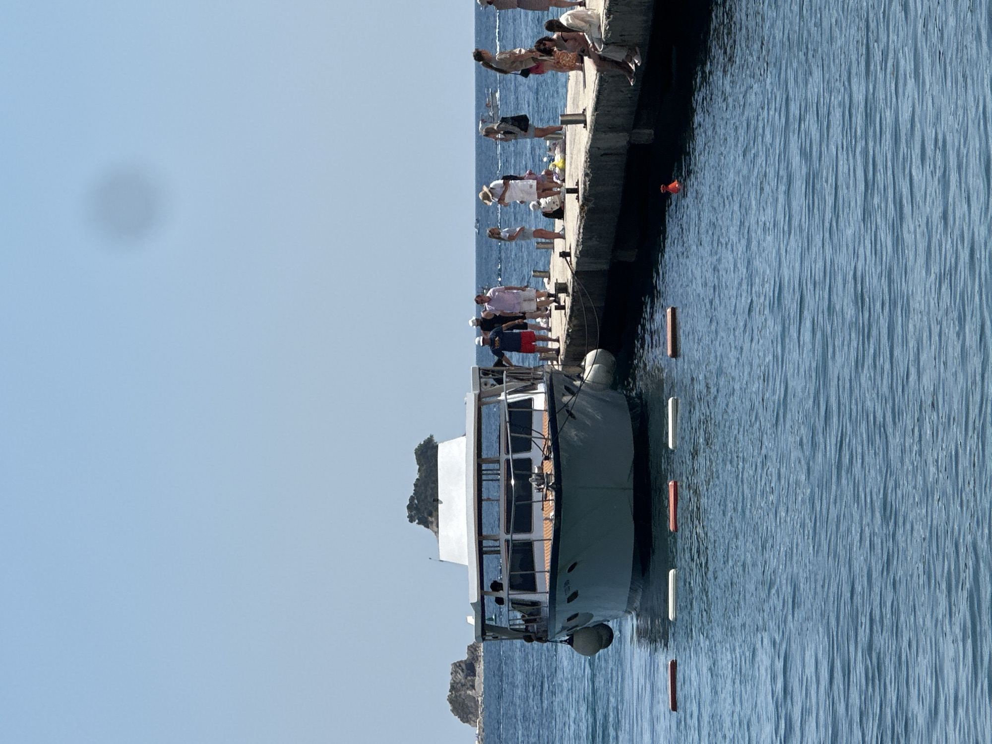 A group of people are gathered on a concrete pier, some standing and some sitting. The pier extends over calm blue water. A white boat with a wooden trim is docked alongside the pier. The boat has several windows and a small deck area. In the background, there are rocky islands with sparse vegetation. The sky is clear and blue, suggesting a sunny day. The people are dressed in casual summer clothing, and some appear to be tourists enjoying the view or waiting for the boat. The overall atmosphere is relaxed and peaceful. There are several buoys floating in the water, some orange and some white.