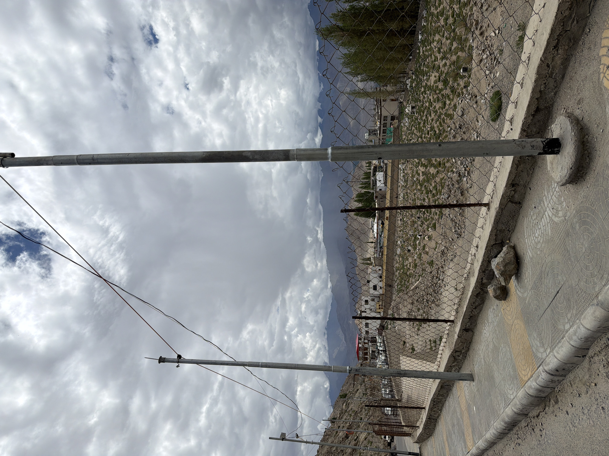 This is a photograph taken in Leh, India, on a cloudy day. The foreground features a paved sidewalk with a curb, bordered by a chain-link fence. Two tall, thin metal poles are positioned along the sidewalk, one closer to the viewer and another further back. The sidewalk is made of concrete tiles with circular patterns, and two rocks are resting on it near the curb. Beyond the fence, there is a sparsely vegetated area with some scattered stones and small buildings. In the middle ground, a street is lined with parked vehicles, and more buildings, some with red roofs, are visible. The background is dominated by a range of imposing mountains under a dramatic, overcast sky with patches of blue visible. The overall impression is one of a slightly rugged, mountainous urban environment.