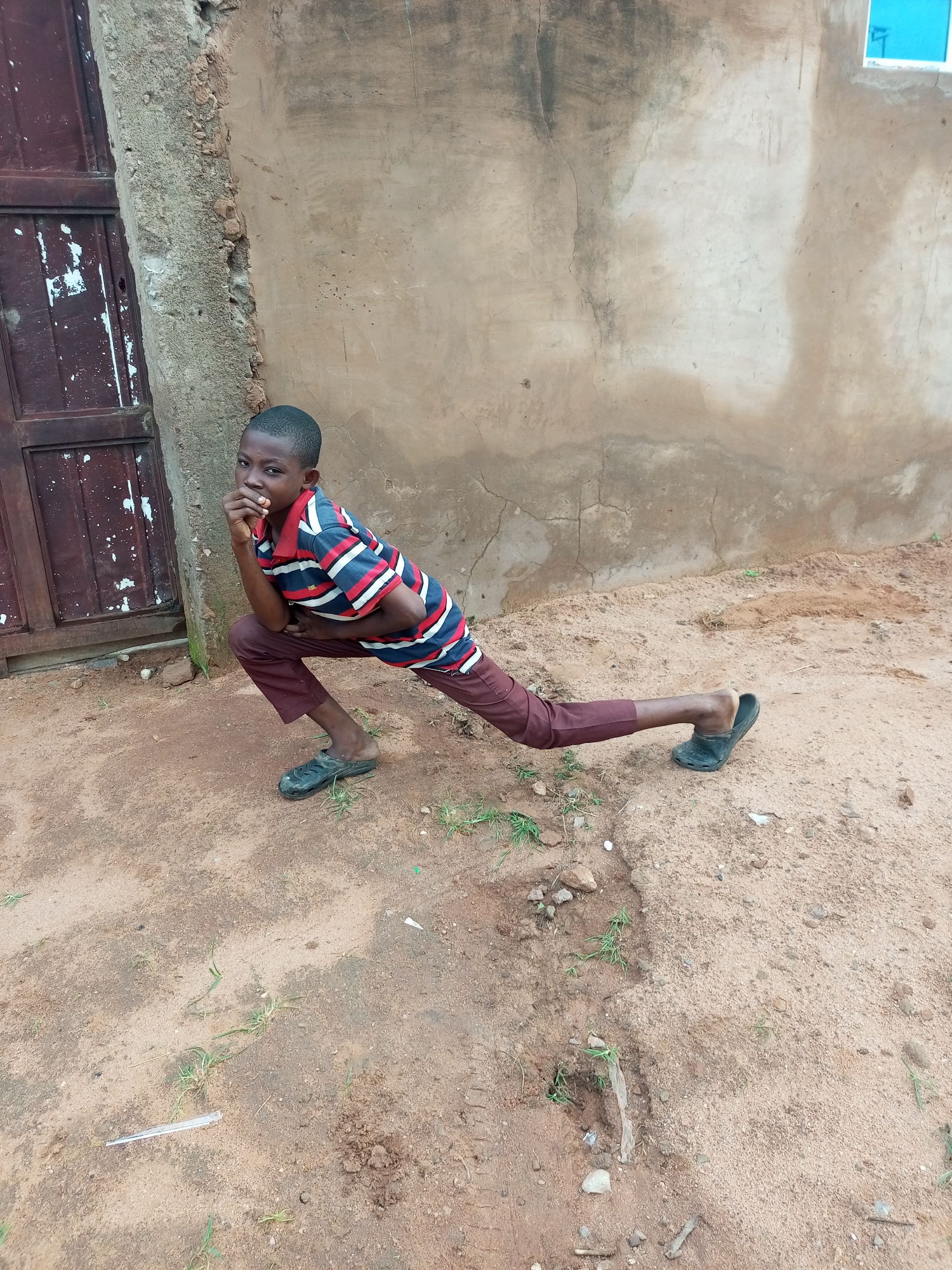 A young boy is posing in a split position in front of a weathered wall and a wooden door. The boy is wearing a striped t-shirt and dark trousers, with black sandals on his feet. He is looking towards the camera with one hand on his chin. The ground is sandy and uneven, with sparse vegetation. A small blue window can be seen on the wall in the upper right corner. The scene appears to be in an outdoor, possibly rural, setting in Tempure, Nigeria.