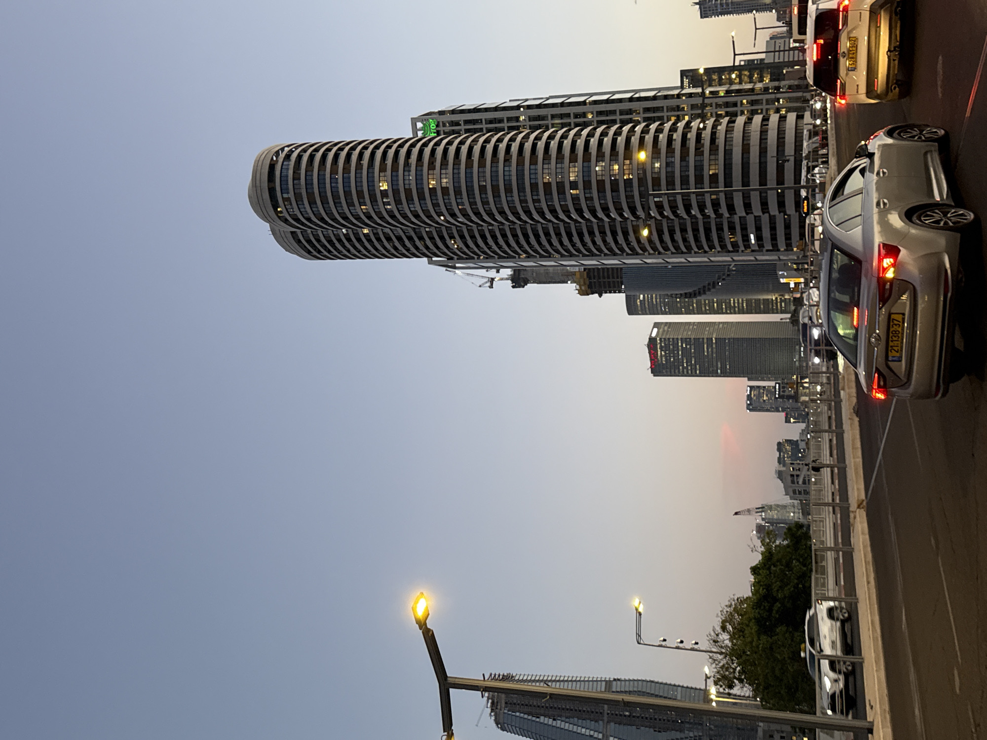 The image depicts a street scene in Tel Aviv, Israel, at dusk. Several tall buildings are visible in the background, including one with a cylindrical, horizontally striped design, likely the Azrieli Center Spiral Tower. The sky transitions from a light blue to a pinkish-orange hue. Several cars are stopped in traffic in the right side of the frame, and their tail lights are illuminated. Street lights line the edge of the road on the right, and modern buildings are visible beyond the streetlights and trees. The image conveys a sense of urban life and cityscape.