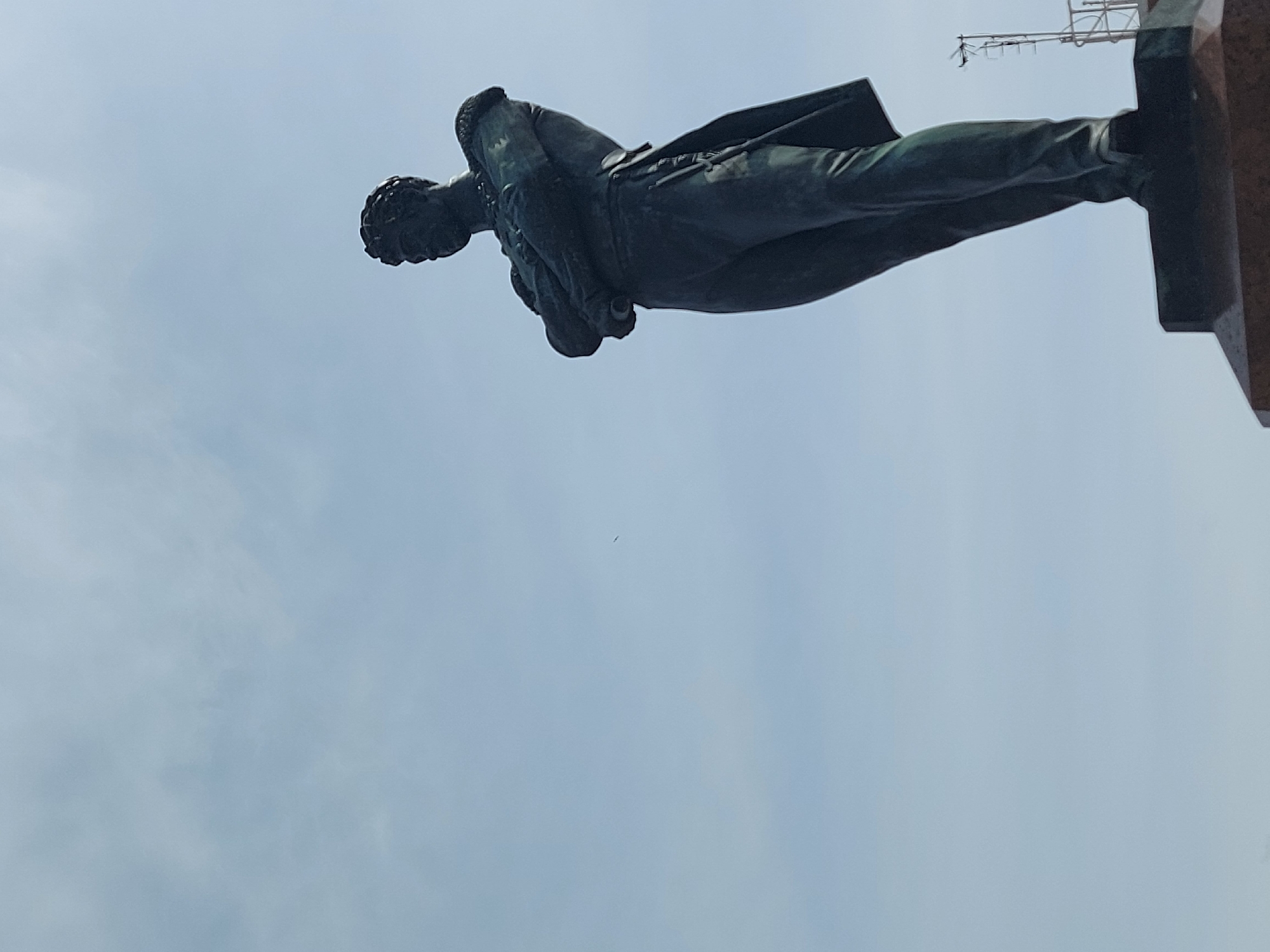The image shows a bronze statue of Vladimir Lenin, viewed from below, against a cloudy sky. The statue stands on a dark, granite base. In the background, some overhead wires and metal structures are visible. This statue is likely the Lenin Monument at Moskovskaya Square in Saint Petersburg, Russia.