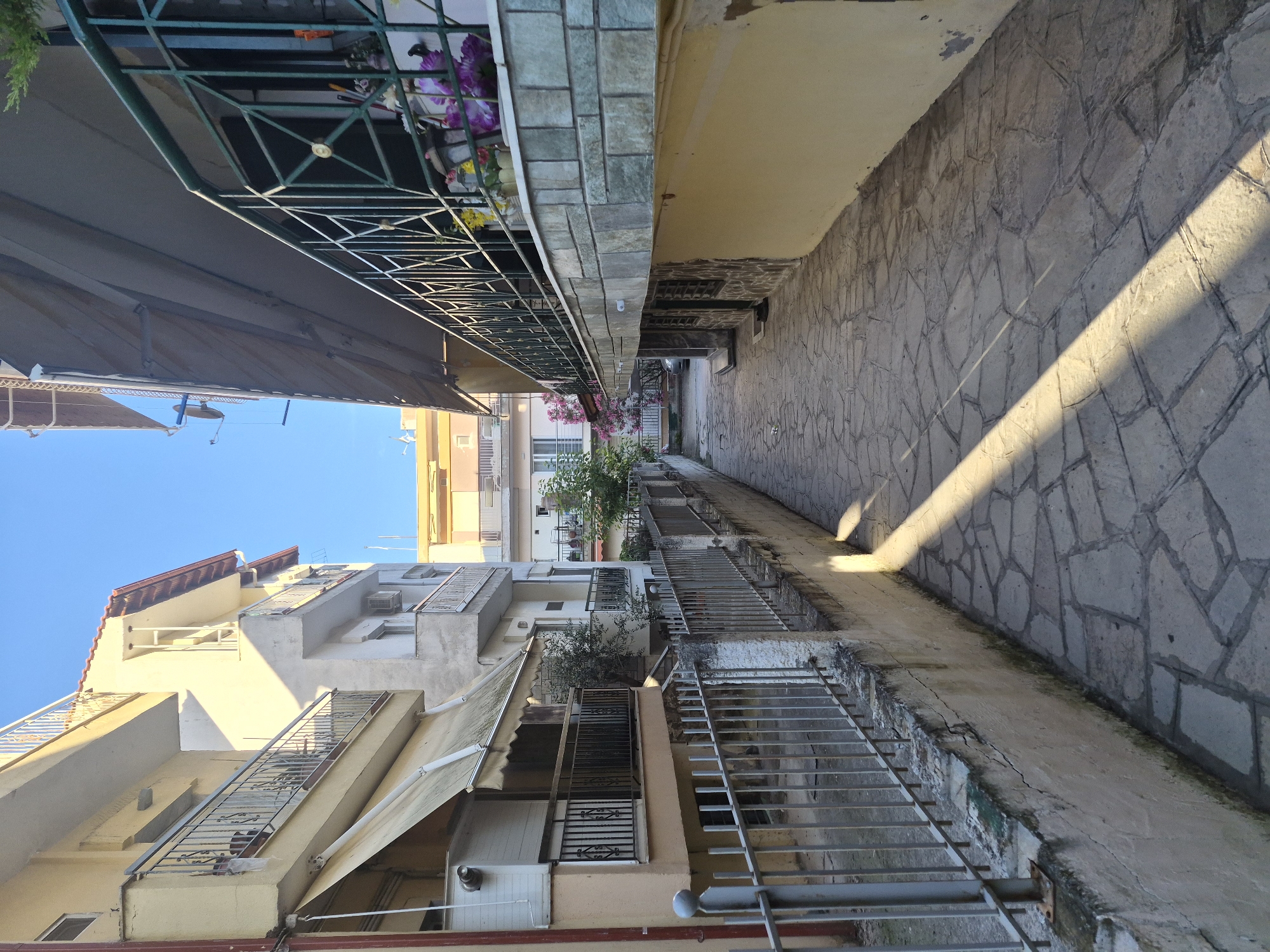 The image depicts a narrow, paved alleyway in the city of Nea Kallikratia, Greece. The alley is bordered by buildings on both sides. On the left, a pale yellow, multi-story building features balconies with metal railings and beige awnings extending over the lower levels. A metal fence runs along the base of this building, separating it from the alley. On the right, a building with a stone-tiled facade rises above the alley, also featuring a balcony with a dark green metal railing adorned with potted plants. The alleyway itself is paved with irregular flagstones, and a diagonal shadow from an unseen object cuts across the pavement. The sky above is clear and blue. The scene appears to be a residential or commercial area, with no people or vehicles immediately visible. The overall impression is one of a sunlit, urban passageway.
