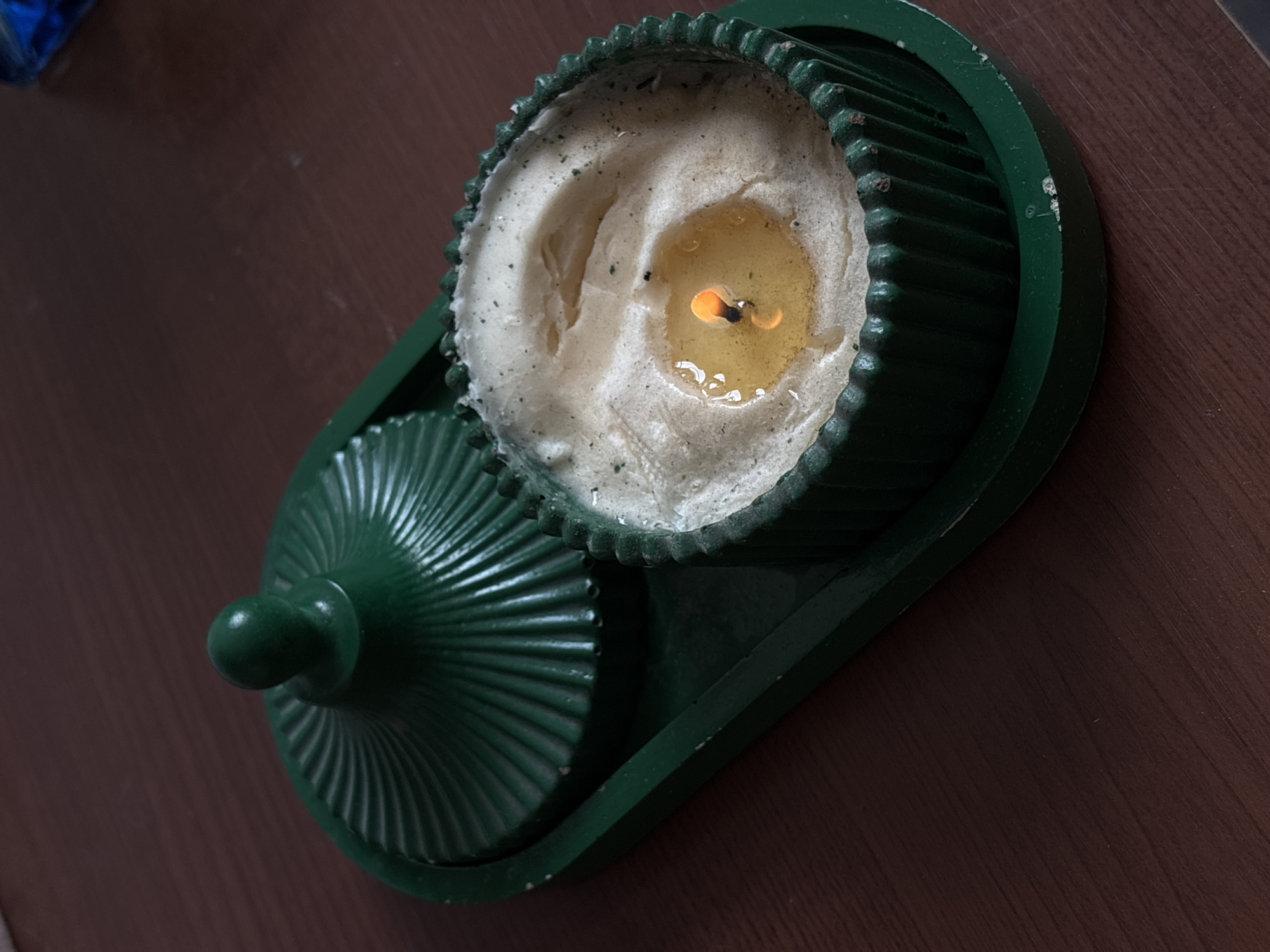 A lit candle rests in a green, lidded container on a dark brown surface. The candle wax is mostly white, with a pool of liquid wax and a visible flame in the center. The container has a circular, ridged base and matching lid with a knob on top. The surrounding surface is a dark brown, possibly wood. The photograph was likely taken indoors in Santa Clara Ocoyucan, Mexico.