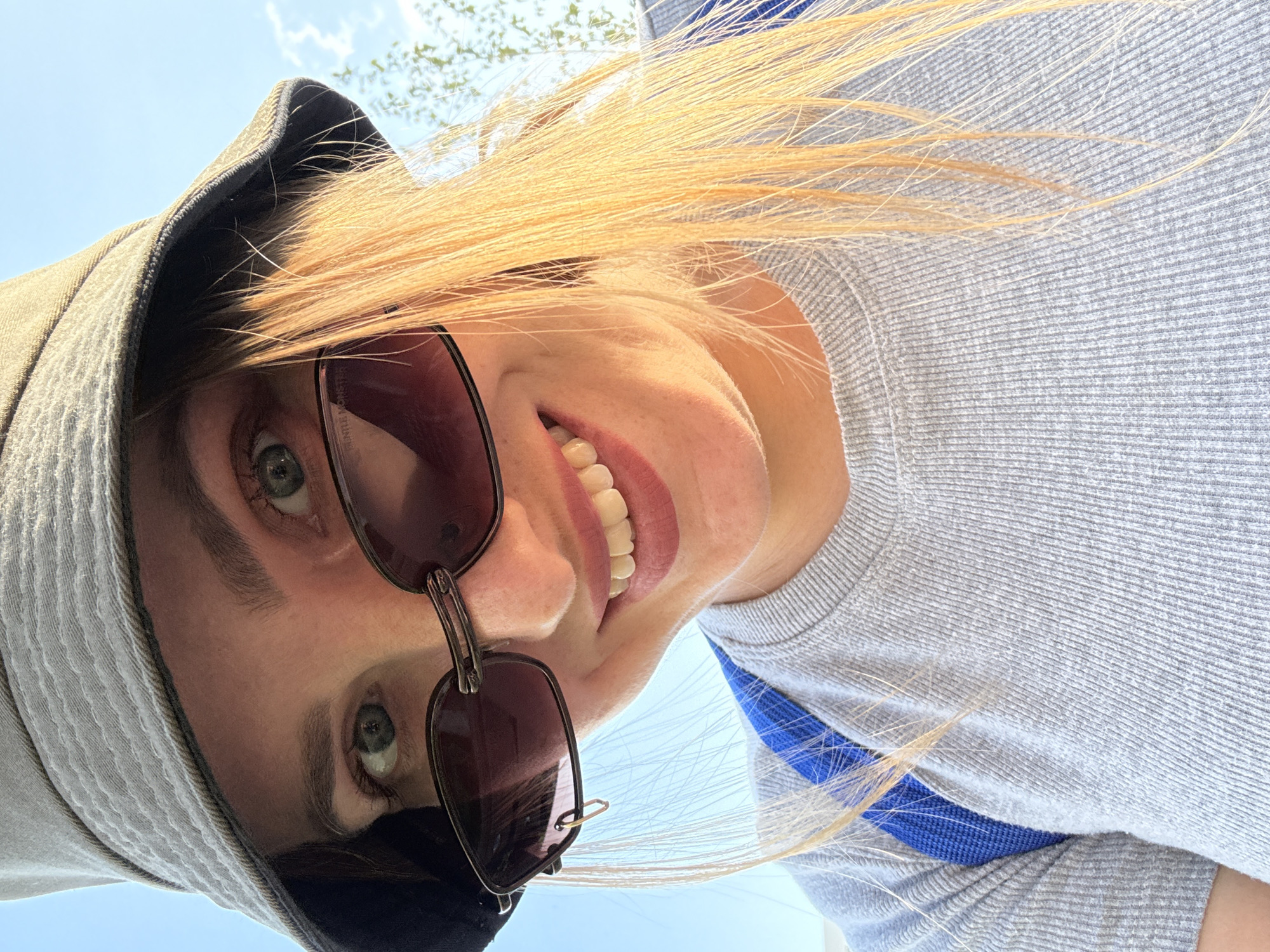 A young woman wearing a grey bucket hat and sunglasses smiles at the camera. She has blonde hair and is wearing a grey ribbed t-shirt with blue straps visible over her shoulders. The background is a clear blue sky with a few wisps of clouds and some blurry greenery. The scene appears to be outdoors, possibly in a park or open area within the city of Kazan, Russia, given the context of the request. There are no other individuals or objects of significance in the frame. The lighting suggests it is daytime, likely sunny.