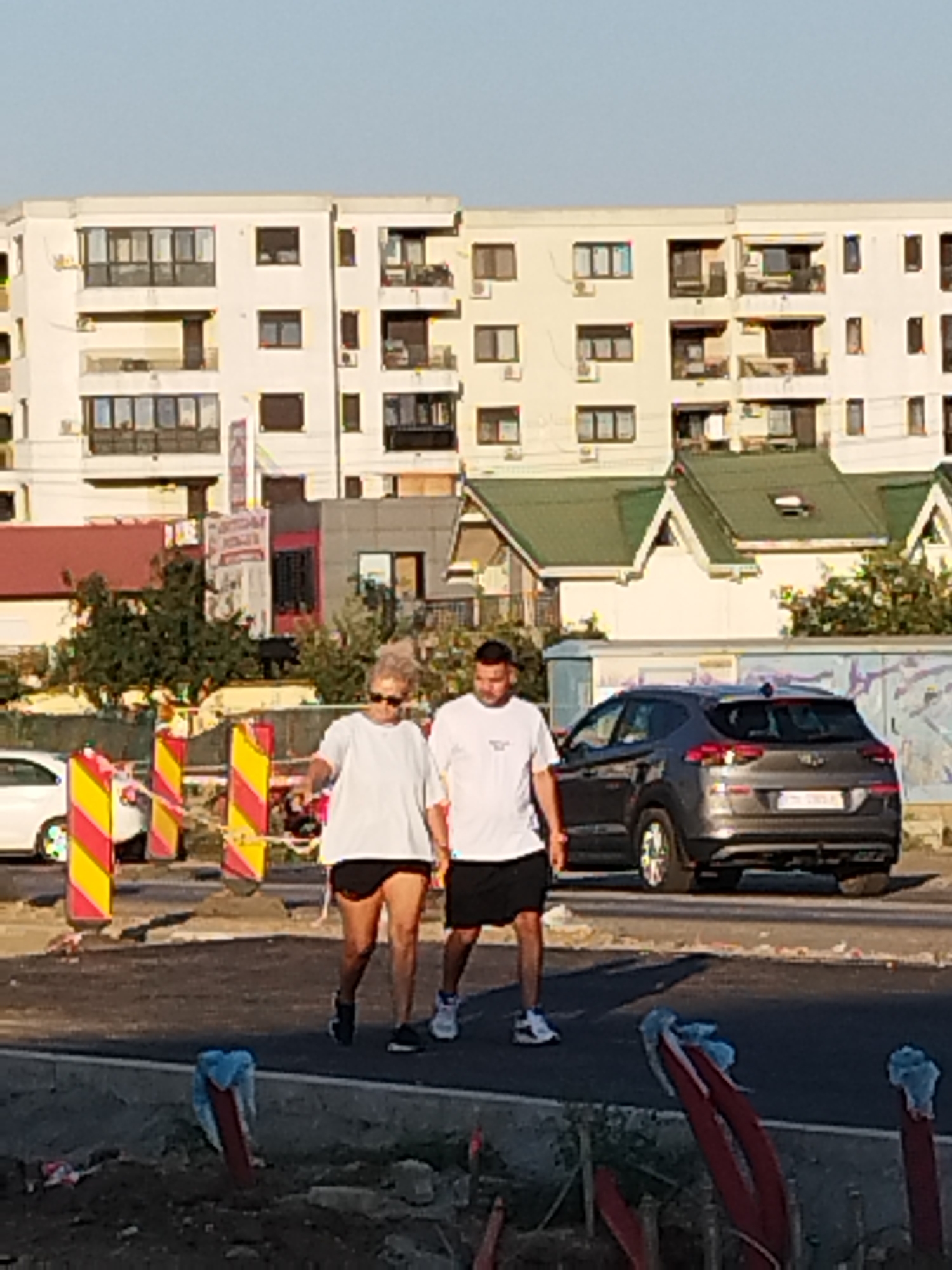 The image captures a daytime scene in Bucharest, Romania, depicting two individuals walking on a street. The man on the right wears a white t-shirt and black shorts, while the woman beside him is in a white t-shirt and black shorts. They are walking towards the left side of the frame, with the man slightly ahead of the woman.
In the background, a modern, multi-story residential building with balconies and numerous windows dominates the skyline. To the right of this building, a structure with a green, sloped roof is visible. A dark-colored SUV is parked on the street to the right of the walking couple.
The foreground shows a construction area with dirt, debris, and several red and yellow striped traffic barriers. Some red pipes wrapped in blue plastic are also present in the foreground, suggesting ongoing urban development or roadwork. The lighting suggests it is late afternoon or early evening, with the sun casting long shadows. The overall environment appears to be an urban setting undergoing renovation.