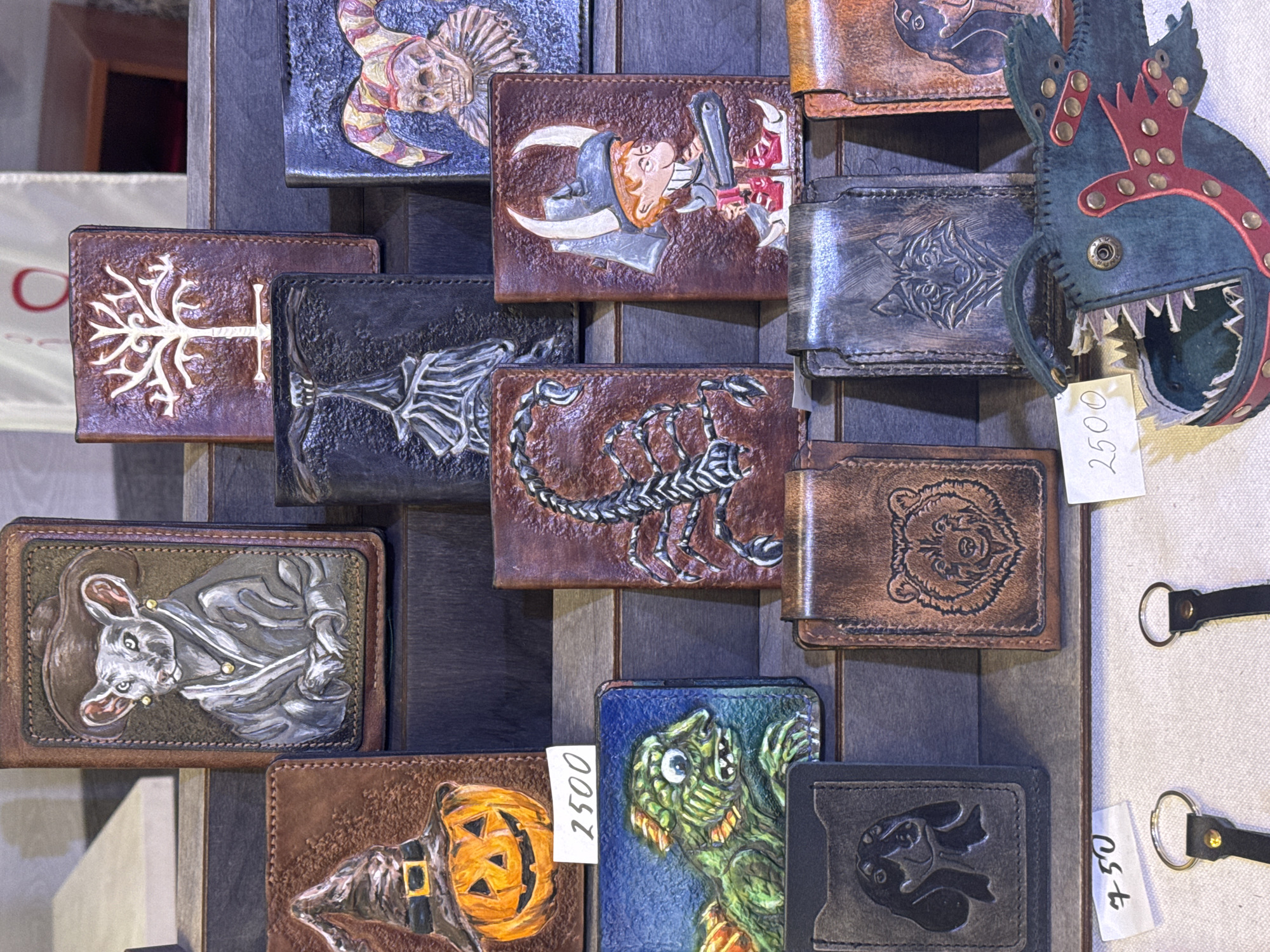A display of hand-tooled leather wallets is presented on a dark-stained wooden rack, possibly within a marketplace or artisan shop. The artwork on the wallets depicts a variety of subjects including fantasy creatures, animals, and Halloween-themed designs. The display is likely situated in Saint Petersburg, Russia, based on the distinctive style of craftsmanship. Several wallets have price tags, suggesting they are for sale. In addition to the wallets, there is a leather key chain shaped like a monstrous fish head. The environment is somewhat shadowed, emphasizing the crafted detail on the leather goods.