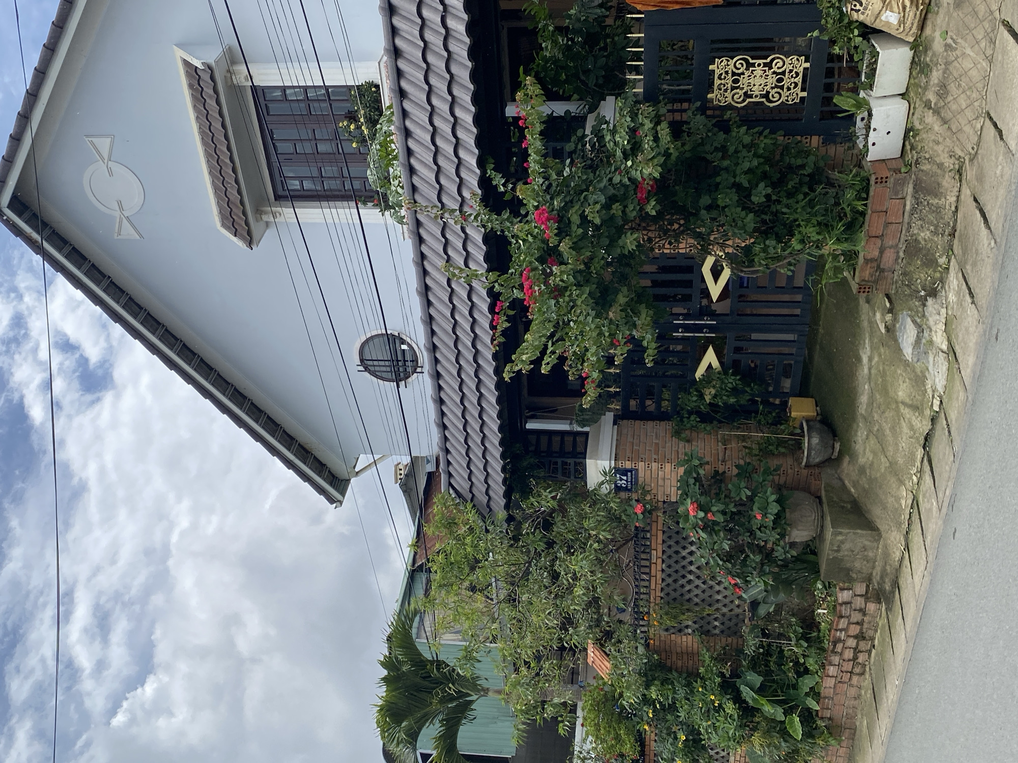 The image depicts a residential property located on Van Hanh street in Da Lat, Lam Vien, Vietnam. The house is a two-story structure with a light blue facade and a dark tiled roof. A prominent feature is a large window on the upper floor with dark brown shutters. Below the upper window, a smaller round window is visible. The front of the house is partially obscured by lush greenery, including flowering bushes with bright pink blossoms and several potted plants. A brick wall with a decorative lattice pattern encloses the property, and a dark metal gate provides access. The street in front of the house is paved and shows signs of wear. The sky is cloudy, suggesting an overcast day. There are no people visible in the image.
