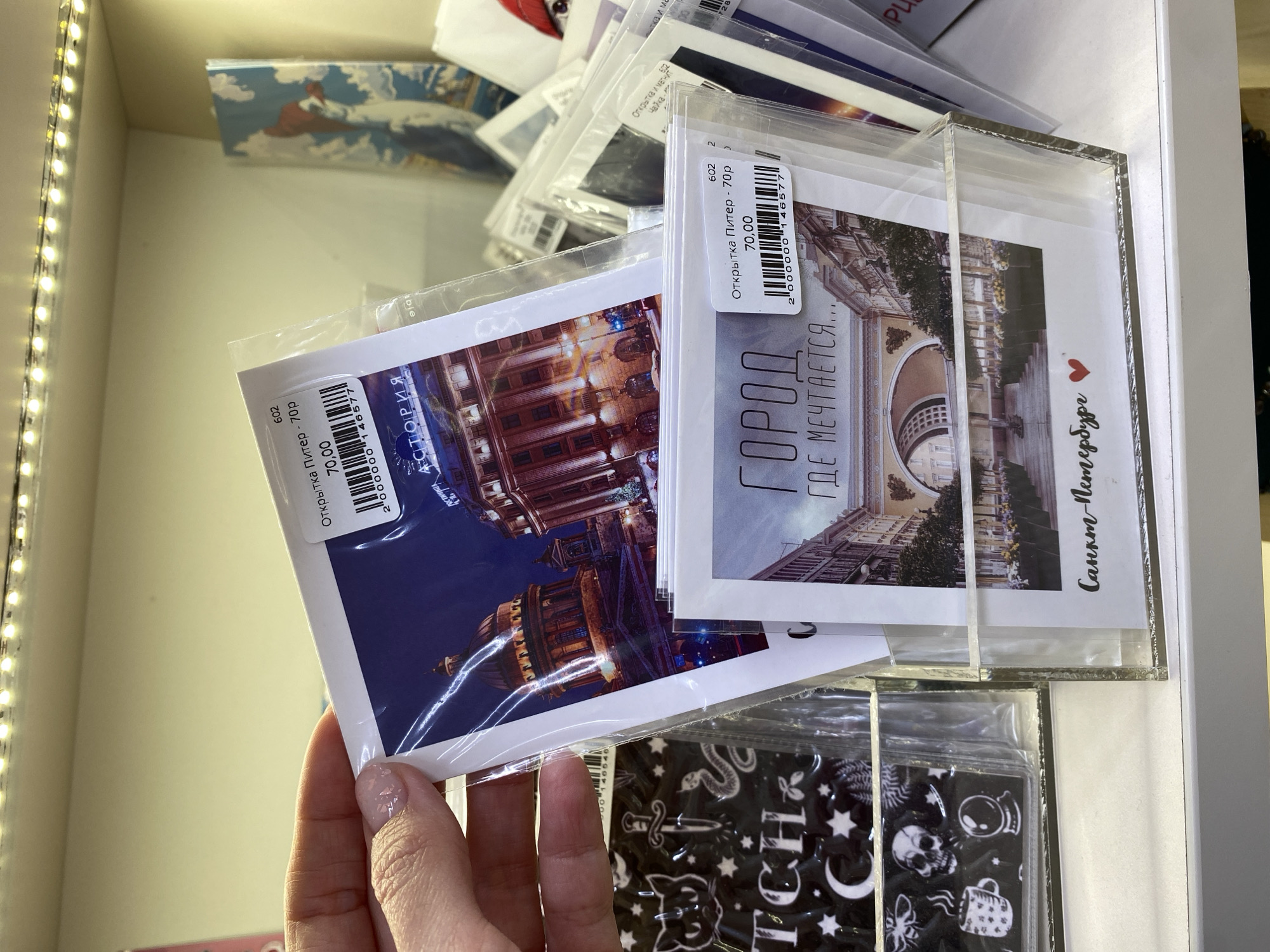 A close-up shot showcases a hand holding a postcard, surrounded by several other postcards in a store setting. The postcard in the hand features a building illuminated at night, possibly a cathedral or palace, with a deep blue sky in the background. To the right, another postcard displays a building during the day, with the text "Город, где мечтается..." ("The city where dreams come true..."). Other postcards in the background include a variety of images, with some featuring illustrations or designs. The postcards appear to be in plastic sleeves and are priced at 70 rubles each. The focus is on the postcards and the hand holding them, with the rest of the store environment blurred.