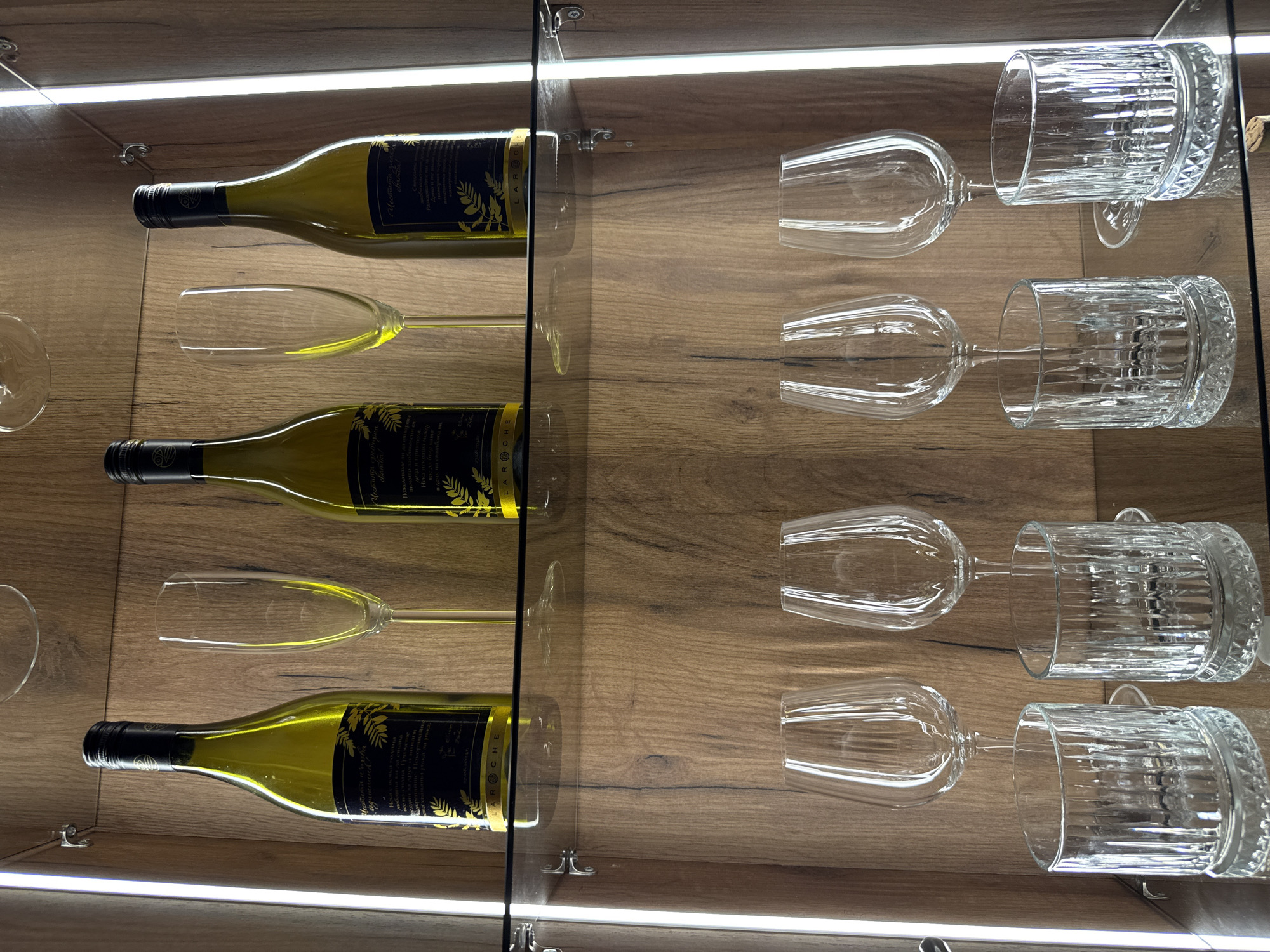 This is a high-angle, eye-level, medium shot of a glass-fronted cabinet displaying alcoholic beverages and glassware. The cabinet has wooden backing and is illuminated by vertical LED strips on the left and right sides.
On the top shelf, three green glass wine bottles with dark labels and black caps are arranged from left to right. Between the first and second bottles, and between the second and third bottles, are two tall, clear champagne flutes with thin stems. The bottles appear to be white wine.
The middle shelf is made of glass.
The bottom shelf, also made of glass, displays glassware. From left to right, there are four glasses: two wine glasses with stems, followed by two shorter, thicker glasses with vertical ridging. All the glassware is clear and empty. A cork is visible on the bottom right edge of the shelf.
The environment appears to be a domestic setting, possibly a home bar or dining area. There is no indication of the location being in the city of Plovdiv, Bulgaria. The scene is static and no people are present.