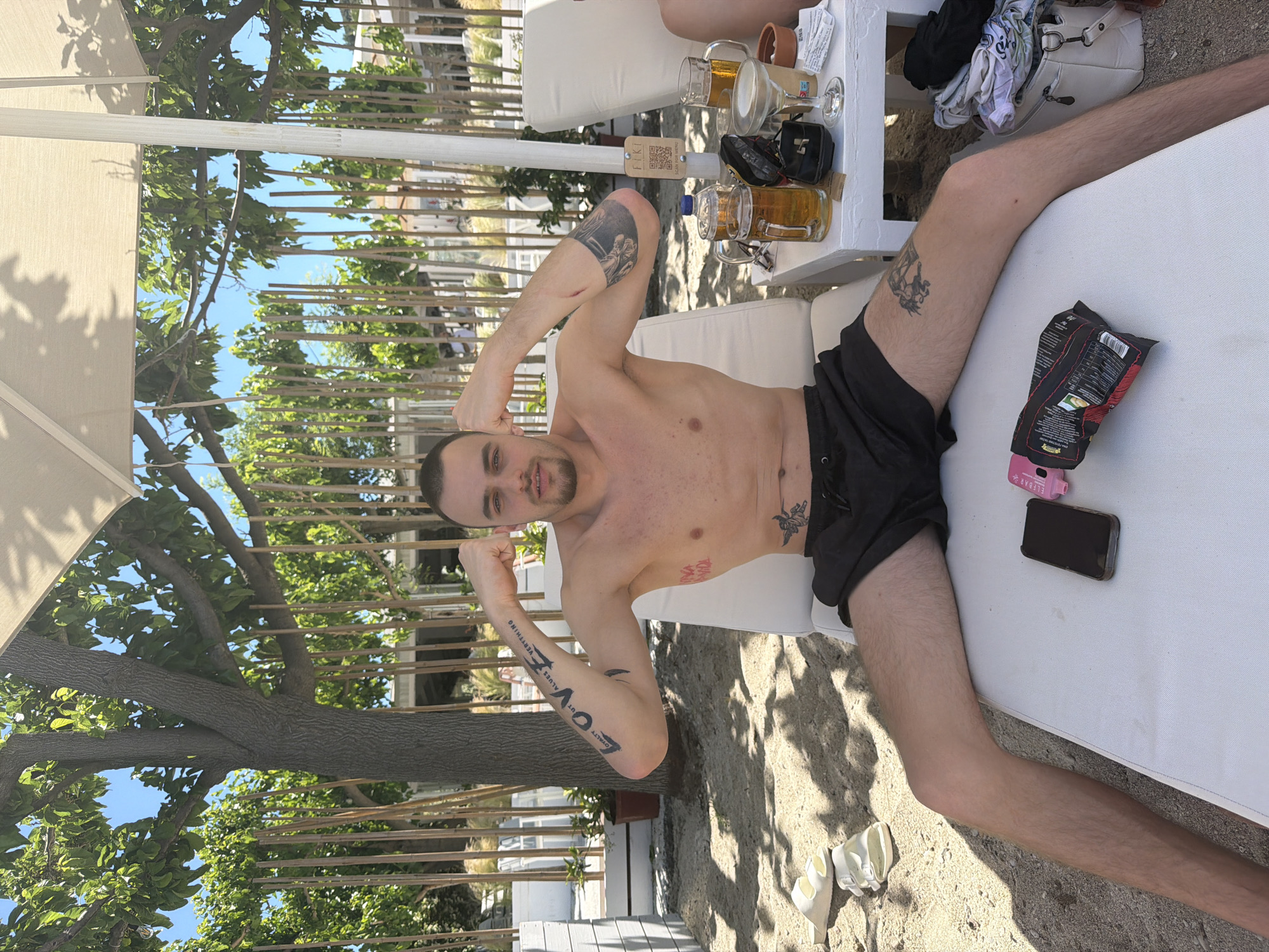 The image shows a man lying on a white cushioned sunbed. He is wearing black swim trunks and has tattoos on his arms, chest, and thigh. He is positioned with his arms raised near his head. On a small white table next to the sunbed are two mugs of beer, a wine glass, a card with the word "FIKI" on it, and some personal items. A bag of crisps and a cell phone are on the sunbed near the man. The scene is outdoor, with sunlight casting shadows on the sunbed and the ground. In the background, there's greenery, a tree, and what appears to be a fence. The image was taken in Pallini Municipal Unit, Greece.