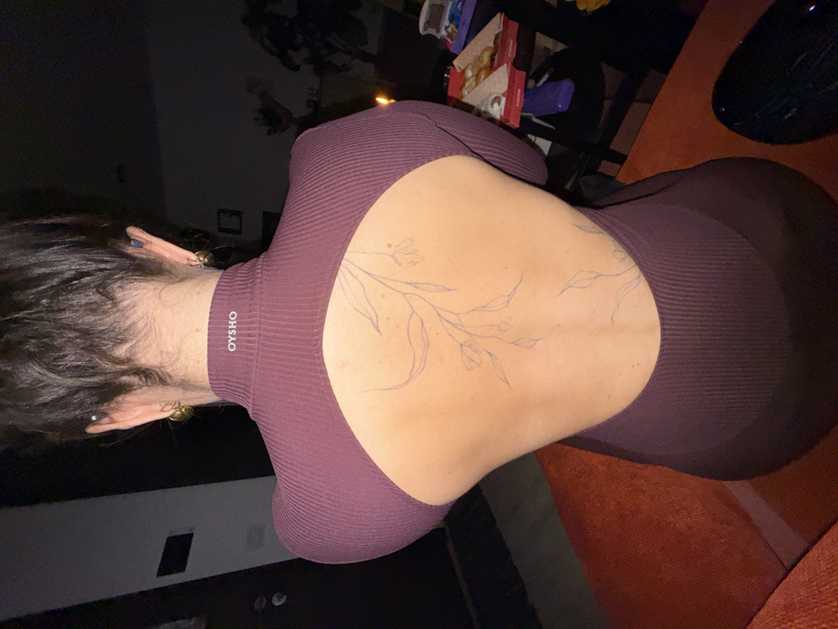 A woman with dark hair, facing away, wears a fitted plum-colored ribbed top with an open back. A floral tattoo design is visible on her bare back. The top has a high neckline with the word "OYSHO" on it. She has earrings in both ears. The background includes a dark table with items on it, a dark wall, and a glimpse of an orange couch. The overall lighting is dim.