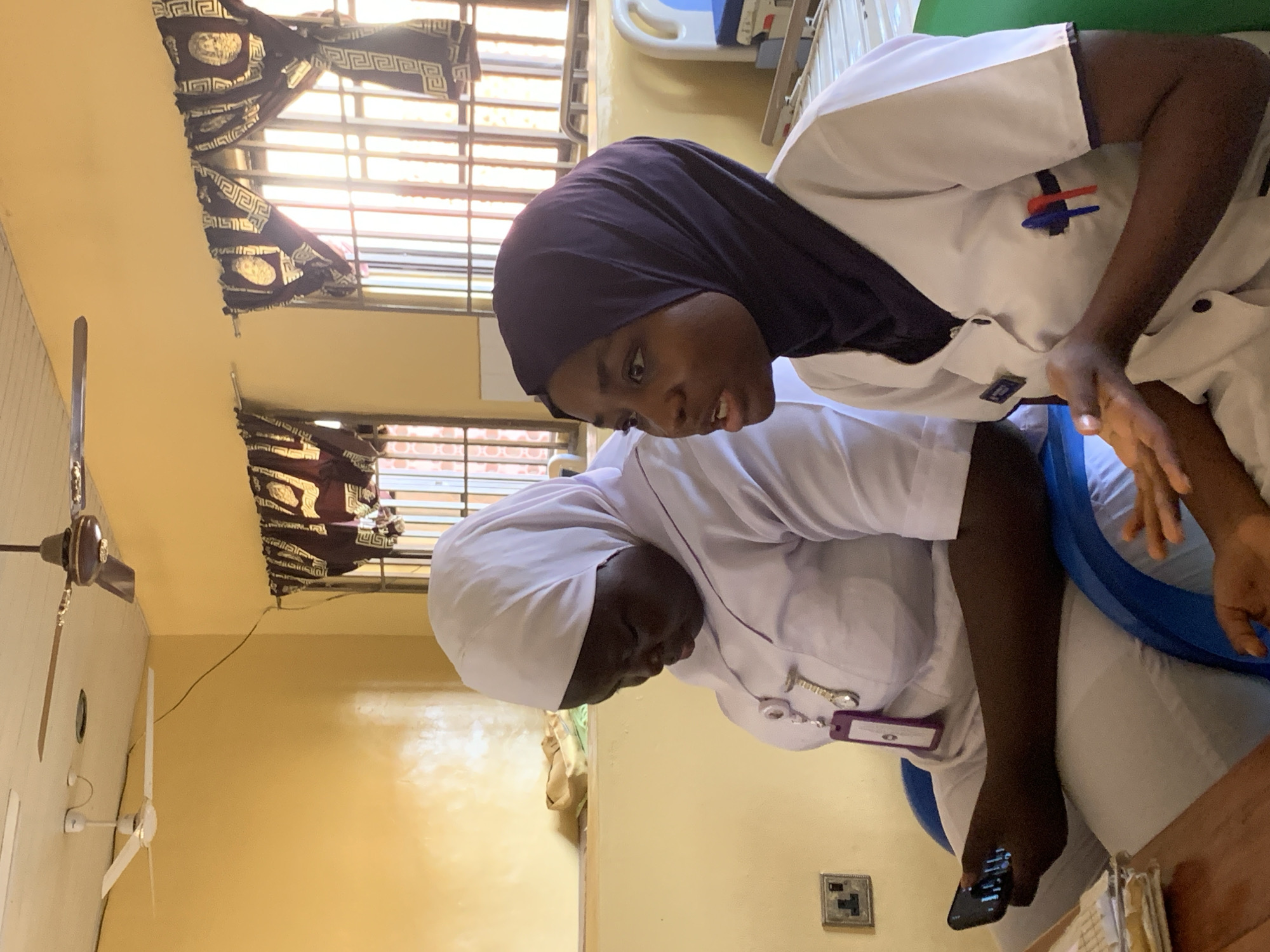 Two individuals are in a medical setting, specifically within a room exhibiting characteristics of a hospital or clinic. One individual is wearing a white hijab and a white uniform. The other individual is also in a white uniform, with a dark head covering. They are looking at something intently. A mobile phone is held by the person in the white hijab. The room has a ceiling fan, and there are curtains on the window. The location is the city of Apata Yakuba, Nigeria.