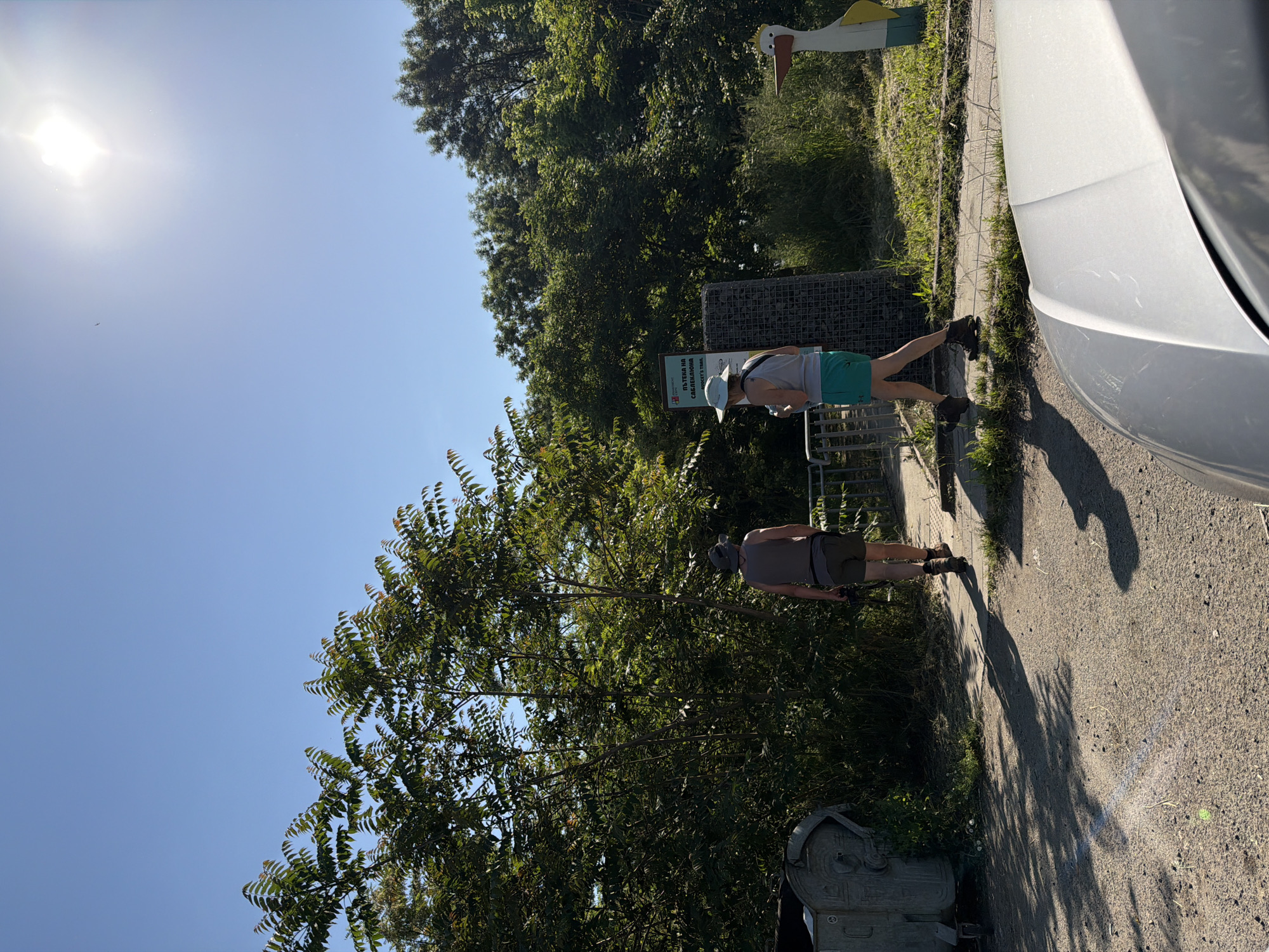 Two people are walking on a paved road next to a car in Burgas, Bulgaria. The person closest to the camera is wearing a grey shirt and brown shorts. The other person is wearing a grey shirt and green shorts, and is looking at a sign. A tall white and yellow bird statue is in the background. There are trees lining the road. The sky is blue with the sun shining.