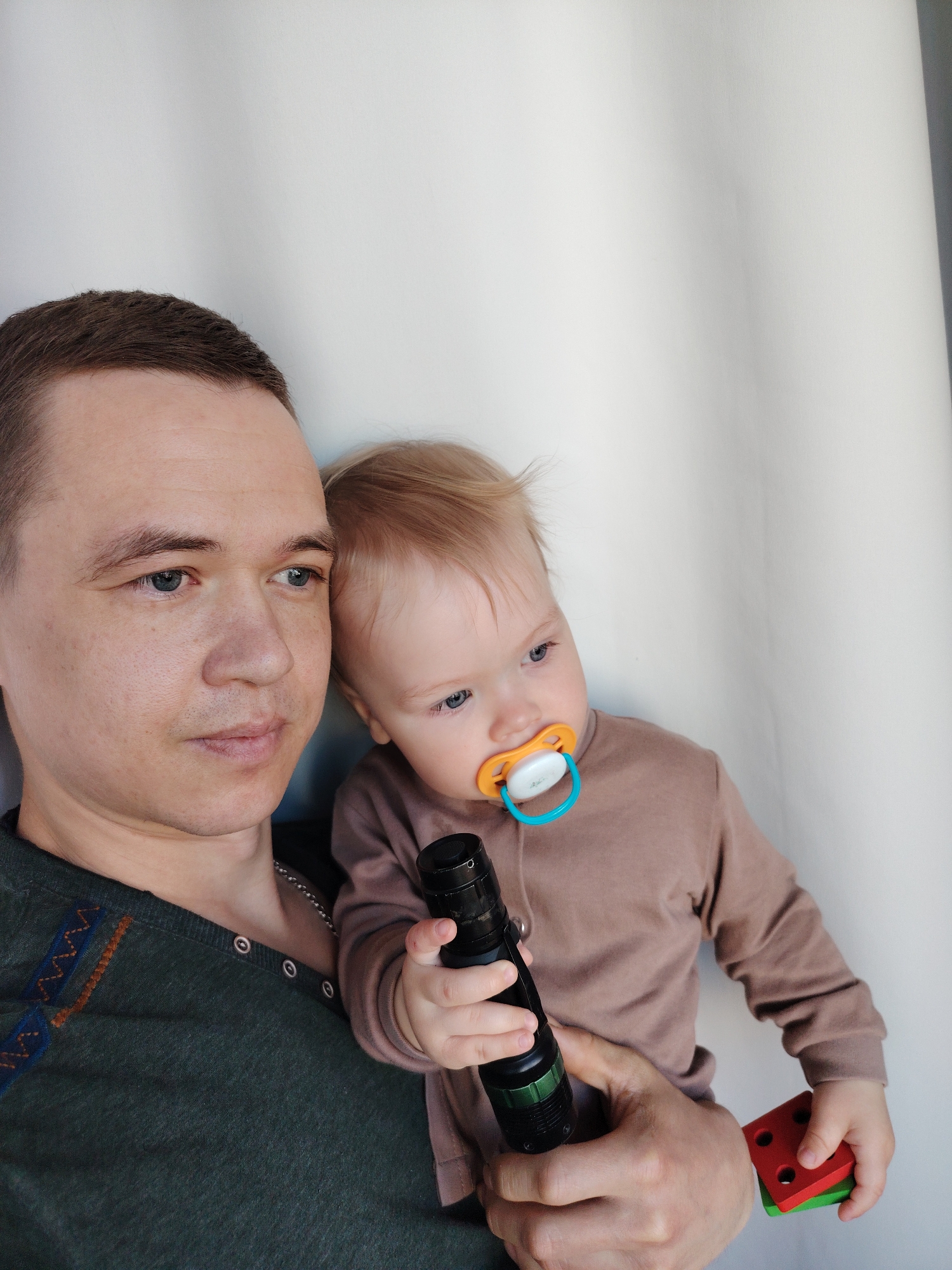 A man is holding a baby with a pacifier in its mouth. The baby is holding a black flashlight in one hand and colorful wooden blocks in the other. The man is wearing a dark green shirt with decorative stitching. The location is Chaikovsky, Russia. The background is a plain white wall. The man and baby are posing indoors.