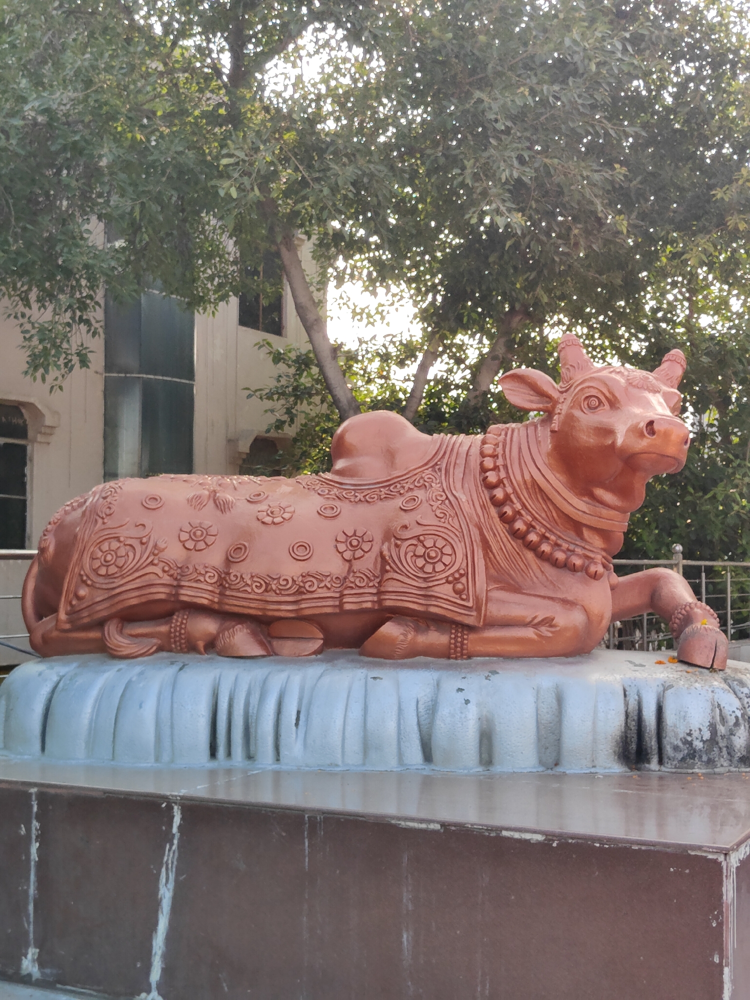 A statue of a cow, likely representing Nandi, the bull of the Hindu deity Shiva, is prominently displayed in the foreground. The statue is orange-brown in color and is adorned with decorative carvings of floral patterns and geometric shapes, suggesting cultural or religious significance. It is seated on a multi-tiered base that transitions from a textured, light-colored upper section with vertical grooves to a darker, marble-like lower section.
The statue and its base are situated outdoors, with a backdrop of trees and a building. The trees, with their dense green foliage, create a natural canopy, and sunlight filters through them, casting dappled light on the scene. The building, partially visible behind the trees, appears to be modern with large windows, one of which has a curved, glass facade. The overall environment suggests a well-maintained public space or religious complex.
The image was taken in Gurgaon, India.