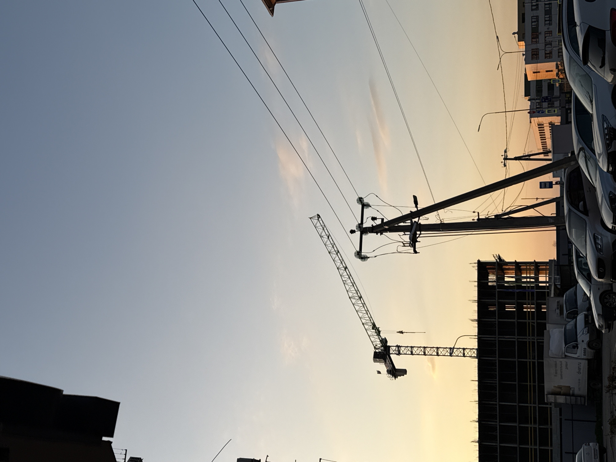 The image depicts a construction site in an urban environment. Two cranes are visible against a sky gradient from pale blue to orange, indicating either sunrise or sunset. A multi-story building under construction is in the background, adjacent to a row of parked cars and trucks. Utility poles with power lines stretch across the sky. Buildings in the distance suggest a city or town. The location is Unknown, Unknown.