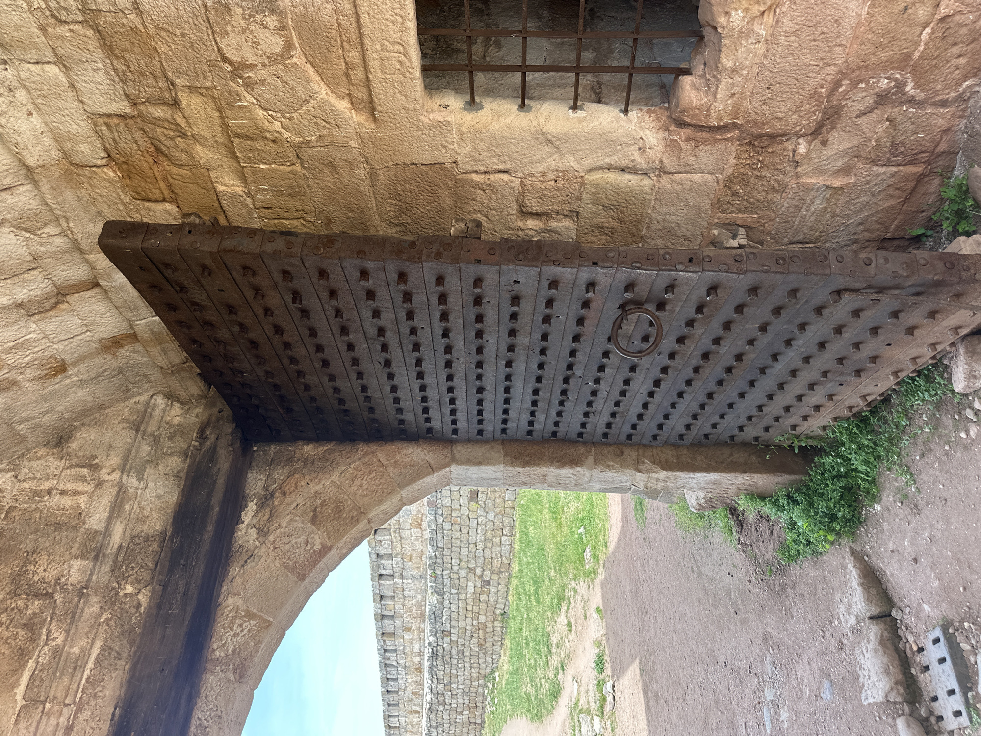 The image shows a heavy, studded iron gate slightly ajar in a stone archway. The location is the Belogradchik Fortress in Belogradchik, Bulgaria. The gate is made of thick planks of wood, reinforced with iron plates and numerous metal studs. The gate features a circular metal handle. The archway is constructed from rough-hewn stones and opens onto a path, grass, and a stone wall. Above the gate, there is a small, barred window in the stone wall. The environment consists of a pathway made of dirt and sand, and overgrown patches of grass.