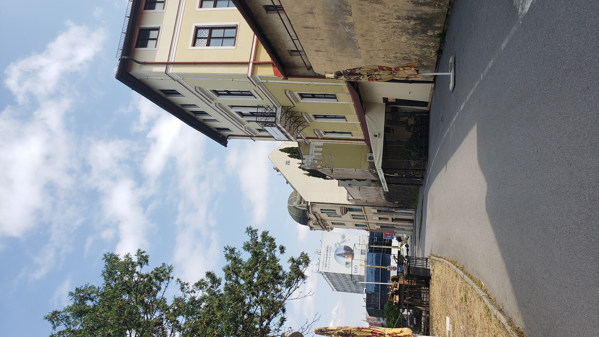 The image shows a street scene in Prešov, Slovakia. On the right side of the street, there is a yellow building with several windows and a balcony. Adjacent to it, a rough, textured beige wall runs along the street. A patterned umbrella stands on the sidewalk in front of this wall. On the left side of the street, a green tree with dense foliage is visible, casting a shadow on the asphalt road. Further down the street, there are several other buildings, including a modern building with a large advertisement billboard and a domed structure. Outdoor seating with tables and chairs is set up on the left side of the road, partially obscured by the tree. The sky is partly cloudy.