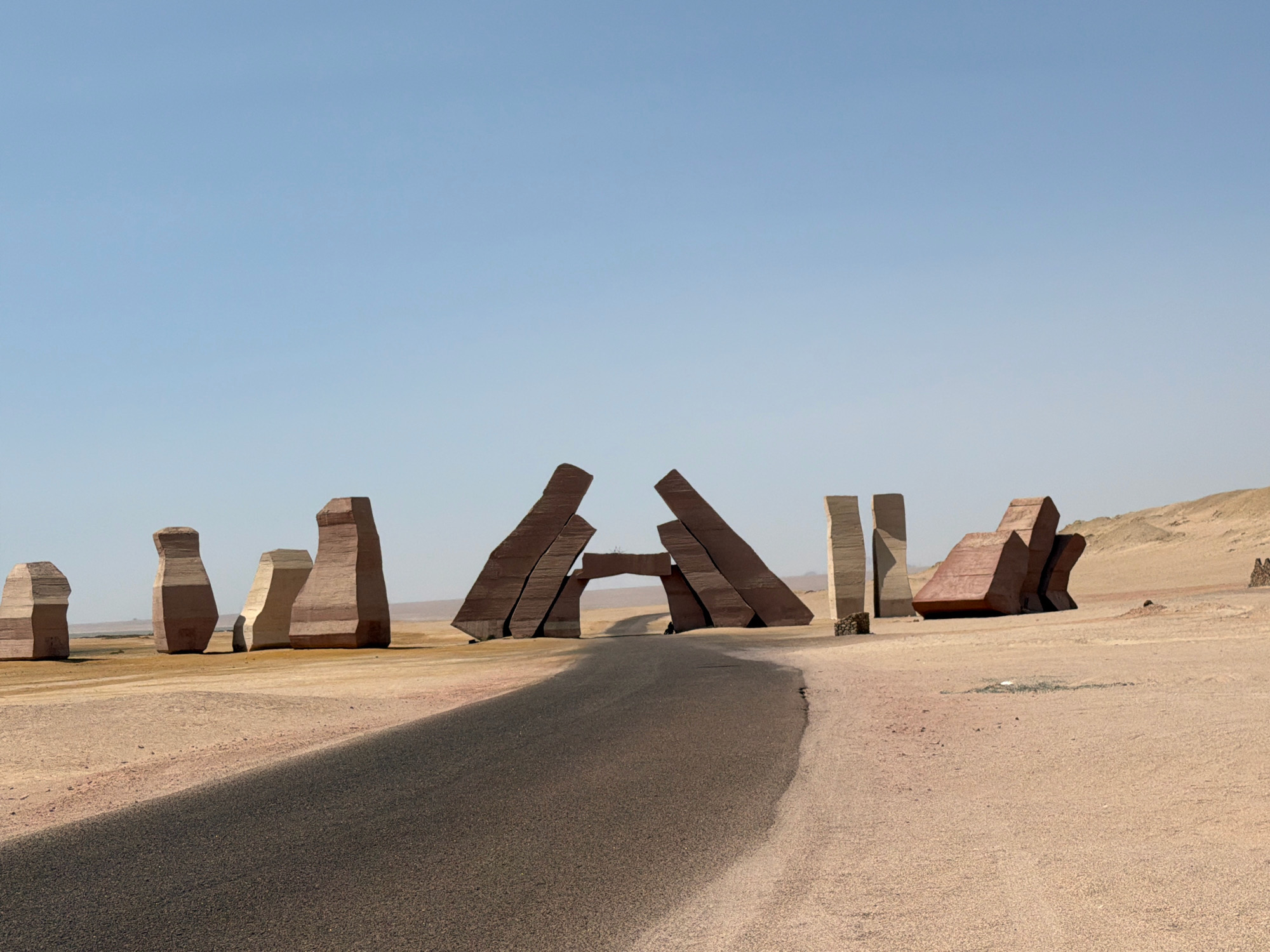 The image depicts a desert landscape in Sharm El Sheikh, Egypt, featuring a series of large, abstract stone sculptures. A paved road curves through the scene, leading towards a prominent archway formed by two tilted, angular monoliths. The sculptures vary in shape and size, with some resembling oversized vases or pillars, and others appearing as sharp, geometric forms. The ground is covered in sand and gravel, with sparse vegetation visible in the distance. The sky is clear and pale blue, suggesting a bright, sunny day. No people are clearly visible in the immediate vicinity of the sculptures or on the road.