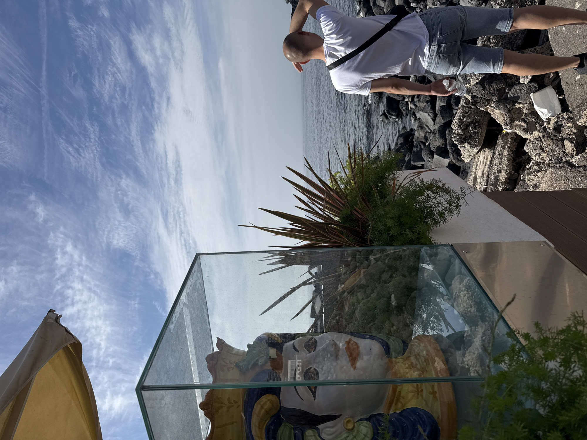 The image captures an outdoor scene in an unidentified location of Unknown, Unknown.
A man, partially visible at the top right, stands near a rocky shoreline facing the sea, clad in a white t-shirt and light blue shorts. He has a dark strap across his chest and holds a clear plastic bottle.
Below, and central to the frame, is a glass-encased ceramic bust. The bust's head is visible, displaying a patterned face with a mustache and a crown or headpiece in hues of yellow, blue, and green. The surrounding landscape is partially reflected in the glass, including the sky, vegetation, and possibly a building. The plants near the bust include greenery and a plant with reddish leaves.
The sky above exhibits blue and white streaky cloud formations. Part of an awning is visible in the bottom left.