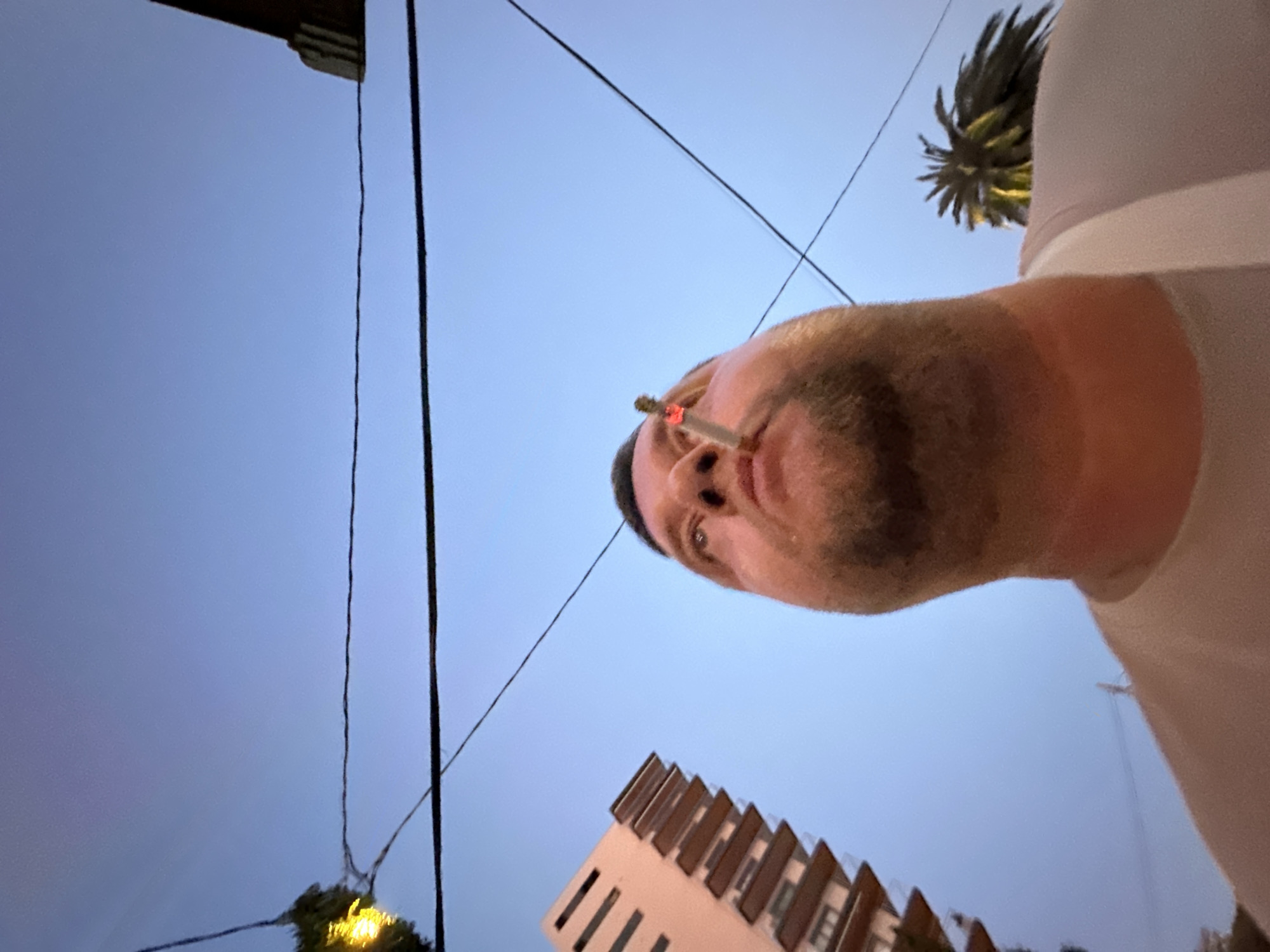 In this low-angle, close-up shot taken in Sant Feliu De Guíxols, Spain, a man is captured looking upwards with a lit cigarette in his mouth. The background reveals a clear blue sky, bisected by several dark power lines. To the left, a portion of a modern building with stacked balconies is visible. To the right, the distinctive fronds of a palm tree peek into the frame. The man is wearing a light-colored t-shirt.