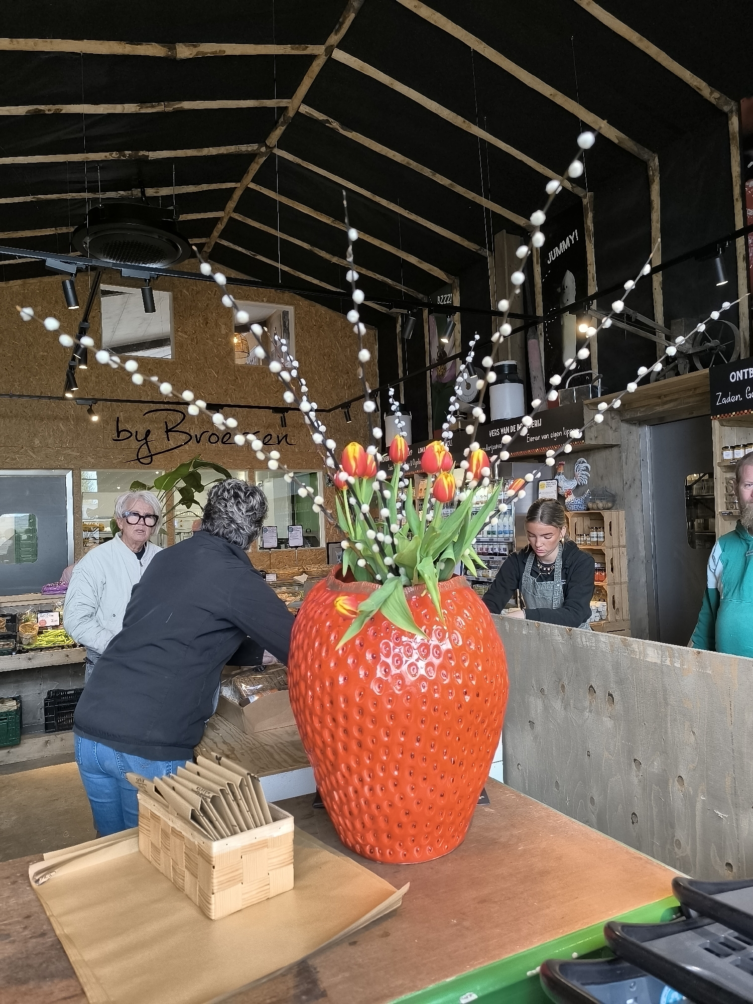 The image captures the interior of what appears to be a retail establishment, possibly a market or store, located in Warmenhuizen, Netherlands. In the foreground, a large, decorative ceramic strawberry acts as a vase, filled with tulips and thin branches adorned with white beads. A small, woven wooden box containing paper sleeves rests on the countertop next to the strawberry.
Two customers are visible near the counter. One is an elderly woman wearing glasses and a light-colored jacket, while the other is a woman with short, gray hair wearing a dark jacket and blue jeans.
Behind the counter stands a young woman, possibly an employee, wearing a dark apron. She appears to be serving or interacting with the customers.
Behind the employee, shelves are stocked with various products. Signage in Dutch is visible, including phrases like "by Broeren," "VERS VAN DE BODERIJ" and "Eieren van elgen kippe." String lights are hung from the ceiling, and there are exposed wooden beams on the ceiling. Further in the background, a man in a green jacket is visible.