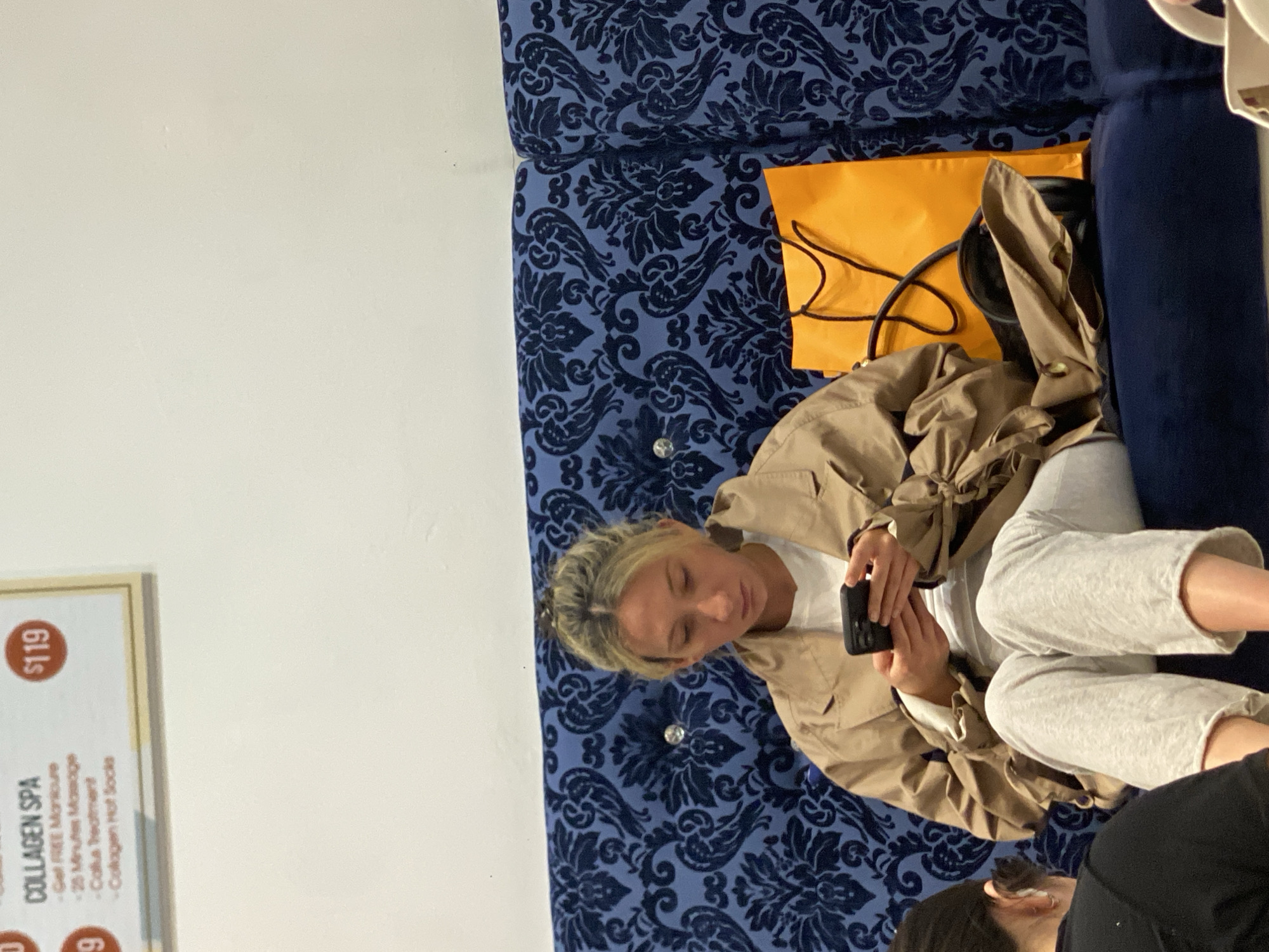A woman is sitting on a blue patterned sofa, looking at her phone. An orange tote bag sits on the sofa next to her. A sign for a "Collagen Spa" is visible on the left side of the frame. The woman is wearing a tan jacket and gray sweatpants. A portion of another person is visible in the lower right corner. The image does not contain enough information to infer the location in New York.