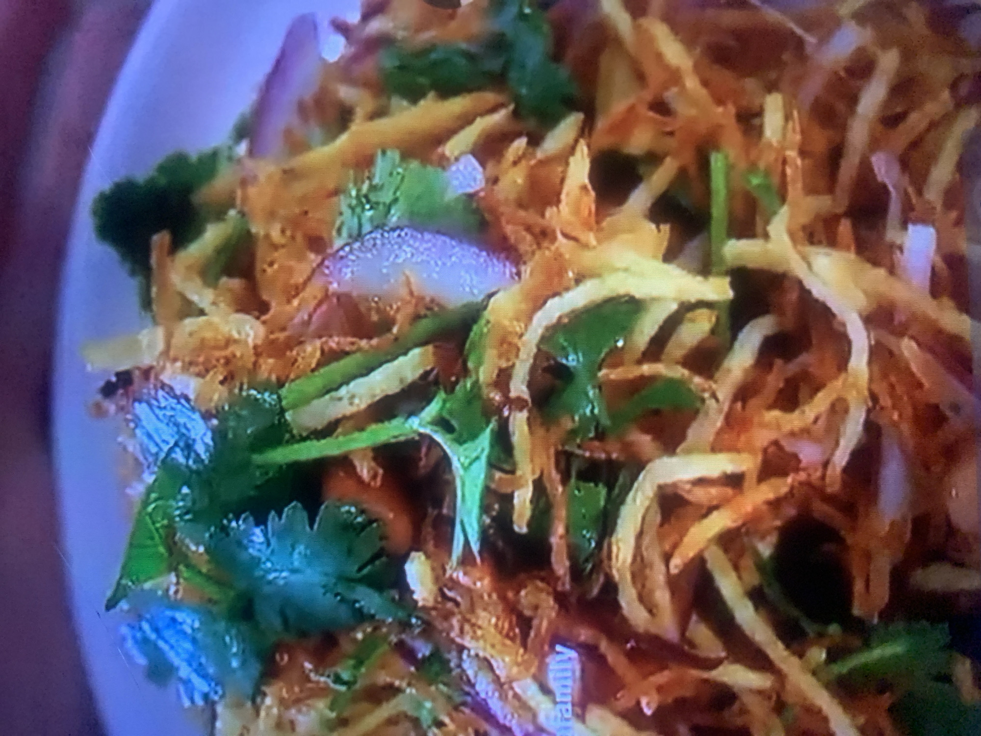 This is a close-up, blurry, and pixelated image of a dish served in a white bowl. The dish appears to be a salad or stir-fry with thin, crispy, golden-brown fried ingredients, possibly noodles or potato shreds, mixed with green herbs like cilantro, and slices of red onion. Some ingredients are green and appear to be leafy or like thin strips of vegetables. The lighting suggests an indoor setting, possibly a restaurant. The overall impression is of a vibrant and flavorful dish.
There are no people visible in the image, and the setting is limited to the bowl of food. The activity is simply the presentation of the food. Notable details include the texture of the fried ingredients and the freshness of the herbs. The image quality makes it difficult to discern specific textures or colors beyond general impressions. There is a faint, partially visible word "family" in white text on a blue background in the bottom right corner of the image, which may be a watermark or part of a logo. No time of day, weather, or emotional cues can be determined from this image. The location context of Almaty, Kazakhstan is provided, but there are no visual cues in the image itself to support this.