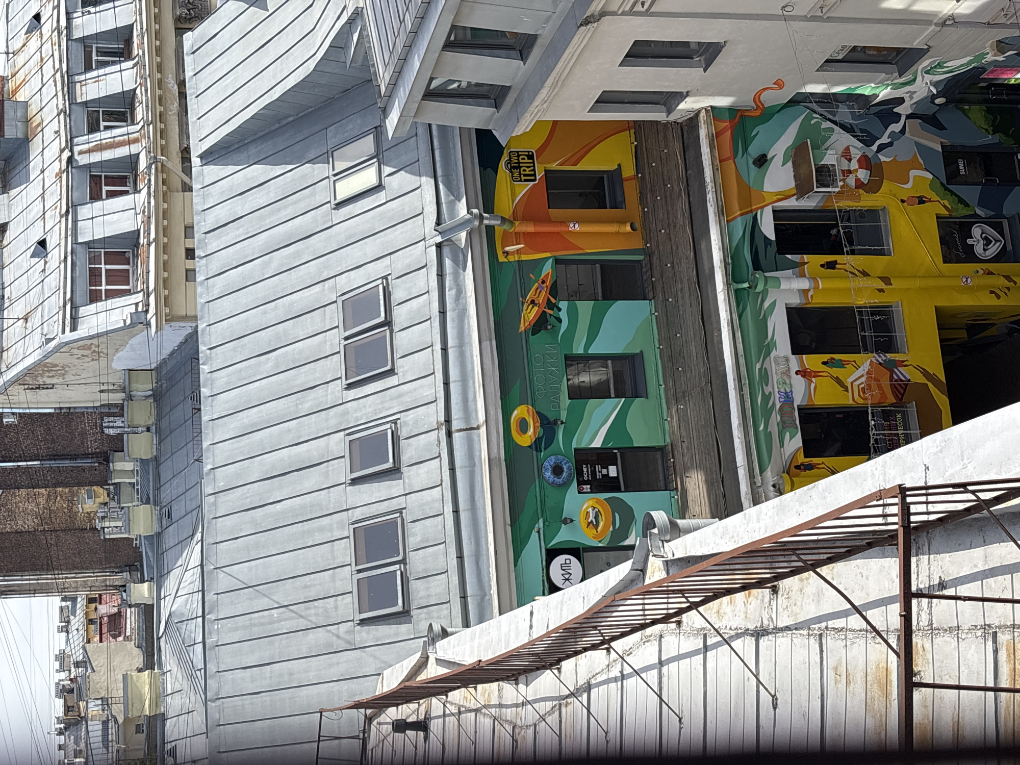 The image depicts an urban scene in Saint Petersburg, Russia, featuring buildings and urban infrastructure. The buildings are predominantly constructed from metal sheeting, exhibiting a gray hue with horizontal seams. One building displays colorful murals, showcasing beach-themed artwork with figures, beach umbrellas, and inflatable rings, and is part of the "One Two Trip" travel company. A rusty metal structure, possibly a fire escape or utility pathway, stretches across the foreground. The lighting is bright, suggesting daytime, and the sky is not visible. Additional buildings are observable in the background, indicating a densely populated urban area.