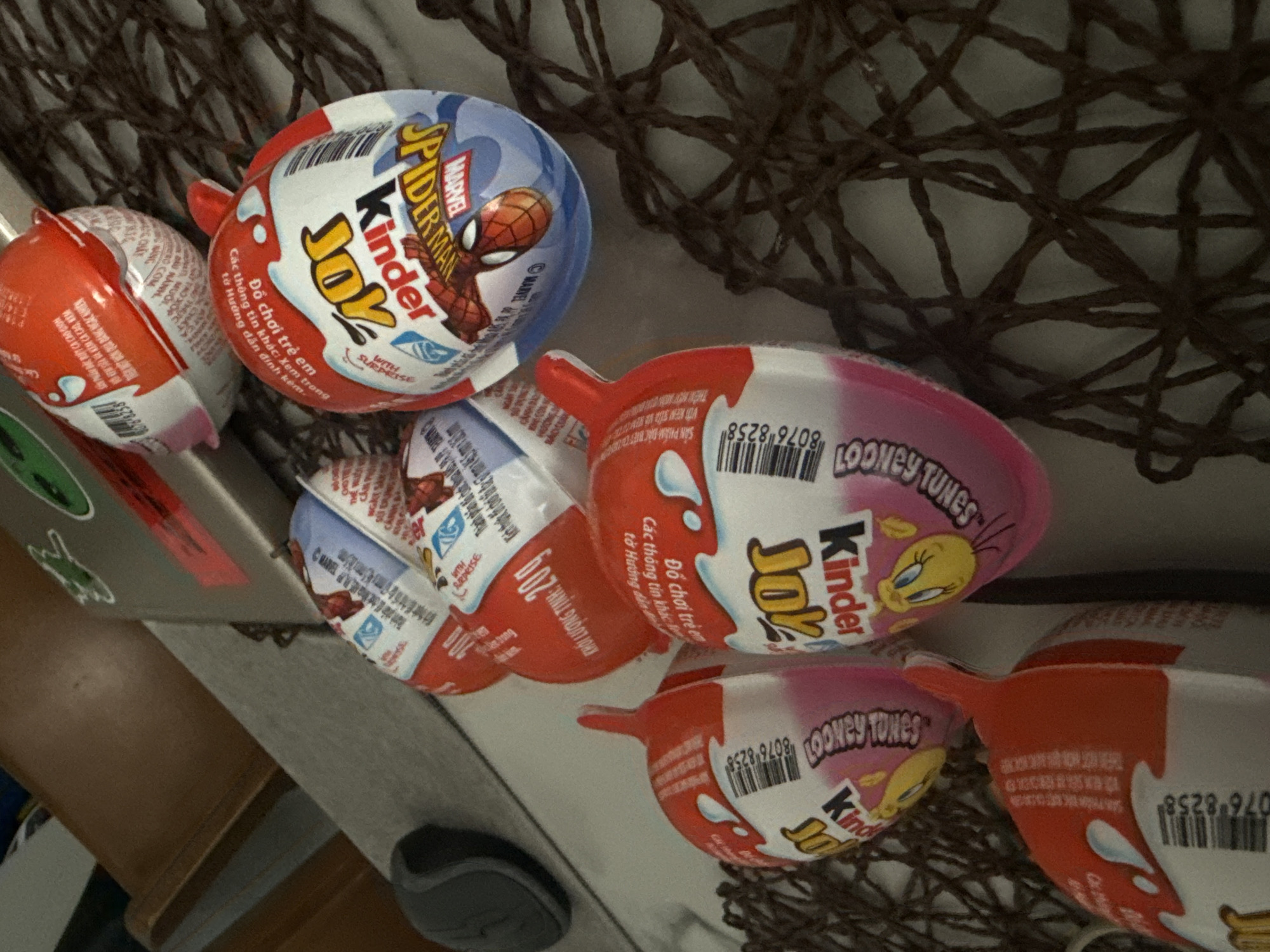 The image shows a collection of Kinder Joy eggs, some with Marvel's Spider-Man branding and others with Looney Tunes characters. These are arranged on a dark, woven mat, likely a placemat. In the background, a laptop is visible with stickers on it, including what appears to be a frog. A dark gray computer mouse is also present. The lighting suggests an indoor setting, possibly a home or cafe. The presence of Vietnamese text on some of the packaging indicates this scene is in Vietnam. While Nha Trang is a city in Vietnam, there are no specific landmarks or contextual clues in the image to confirm its precise location within the city.
