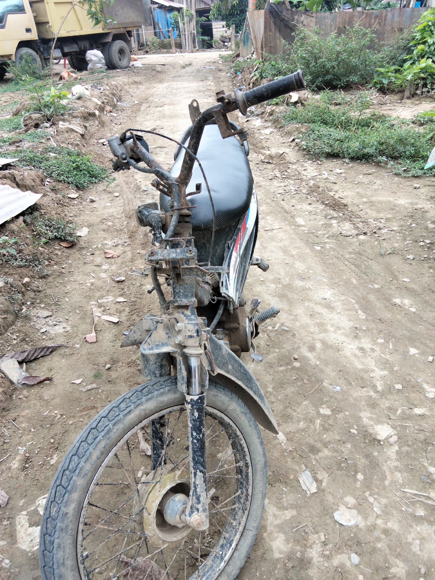 The image shows a damaged motorcycle on a dirt road in Benuaraja, Indonesia. The motorcycle is black with some red and white accents. It appears to be old and weathered, with rust on the handlebars and front forks. The front wheel is visible, and it is dirty. The road is unpaved and appears to be in disrepair, with loose dirt and small rocks. In the background, there is a yellow truck, some vegetation, and buildings that appear to be houses. The scene is likely a residential area or a rural part of the city. There are no people visible in the image. The lighting suggests it is daytime.