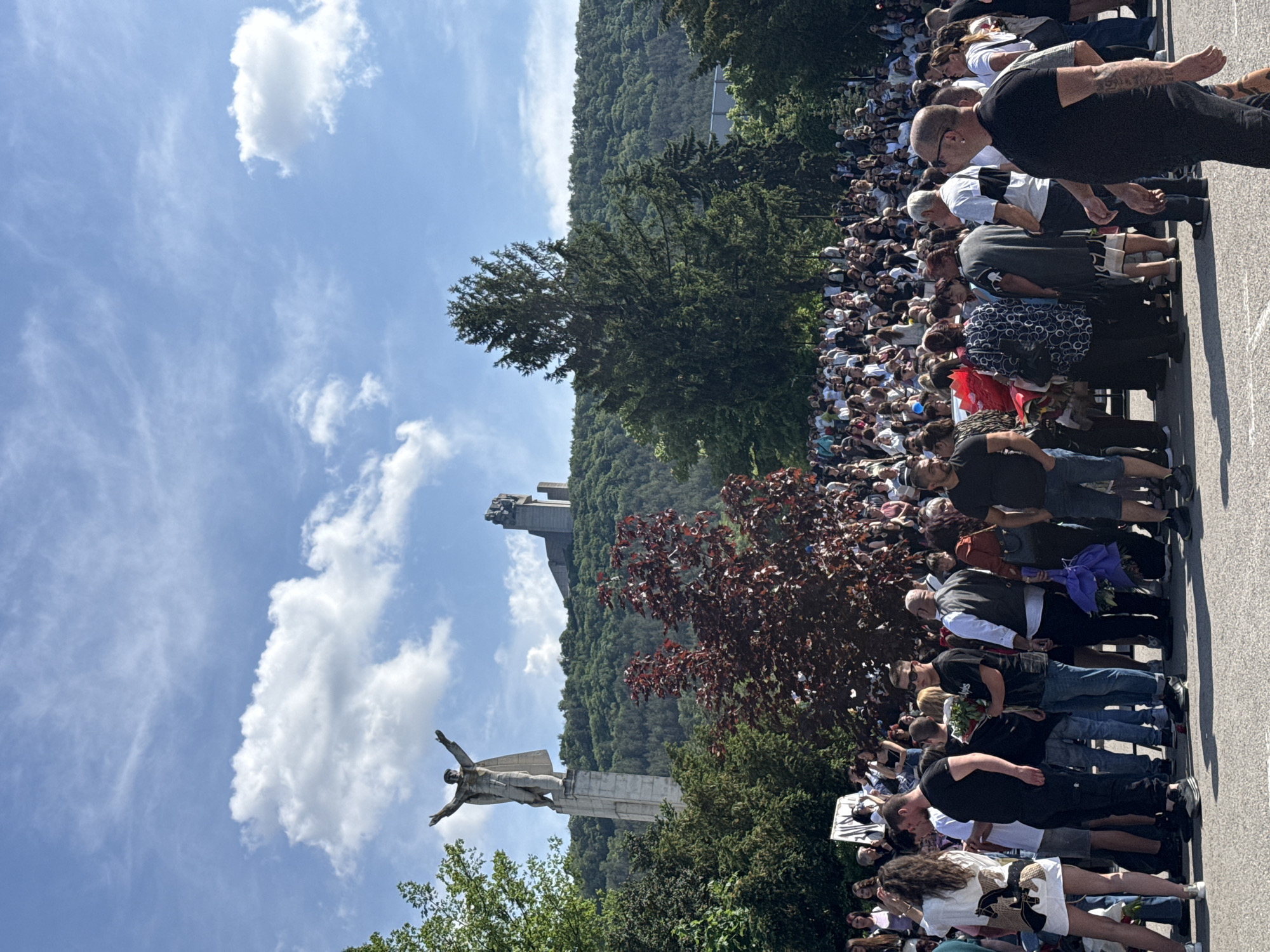 The image shows a large crowd of people gathered at the Founders of the Bulgarian State Monument in Shumen, Bulgaria. The monument is a prominent landmark featuring a winged statue and other sculptural elements. The crowd is densely packed, suggesting a public event or gathering. The sky is blue with scattered clouds. The monument and surrounding area are situated on a hilltop, surrounded by trees. People are standing and observing the monument. Some may be holding flowers.