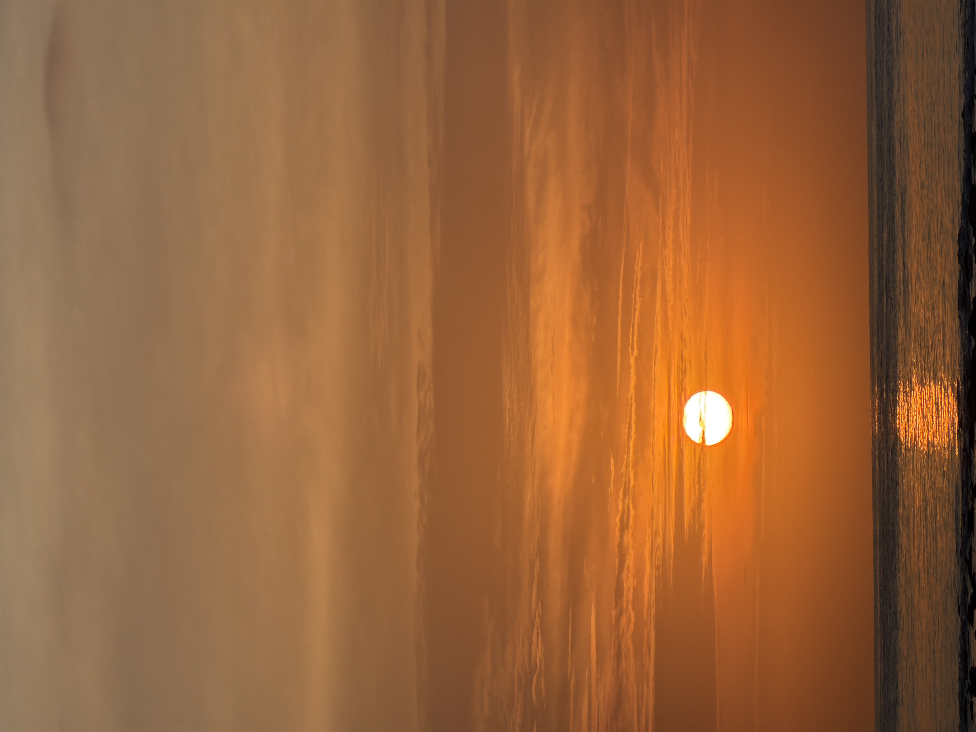 The image depicts a serene sunset over the ocean. The sun, a bright, glowing orb, is positioned slightly to the left of the center, casting a warm, orange hue across the sky and reflecting on the water's surface. Wispy clouds are scattered throughout the sky, adding texture to the gradient of orange and yellow. The ocean is calm, with gentle waves lapping at the shore. The foreground shows a rocky shoreline. The location is identified as Unknown, Indonesia.
