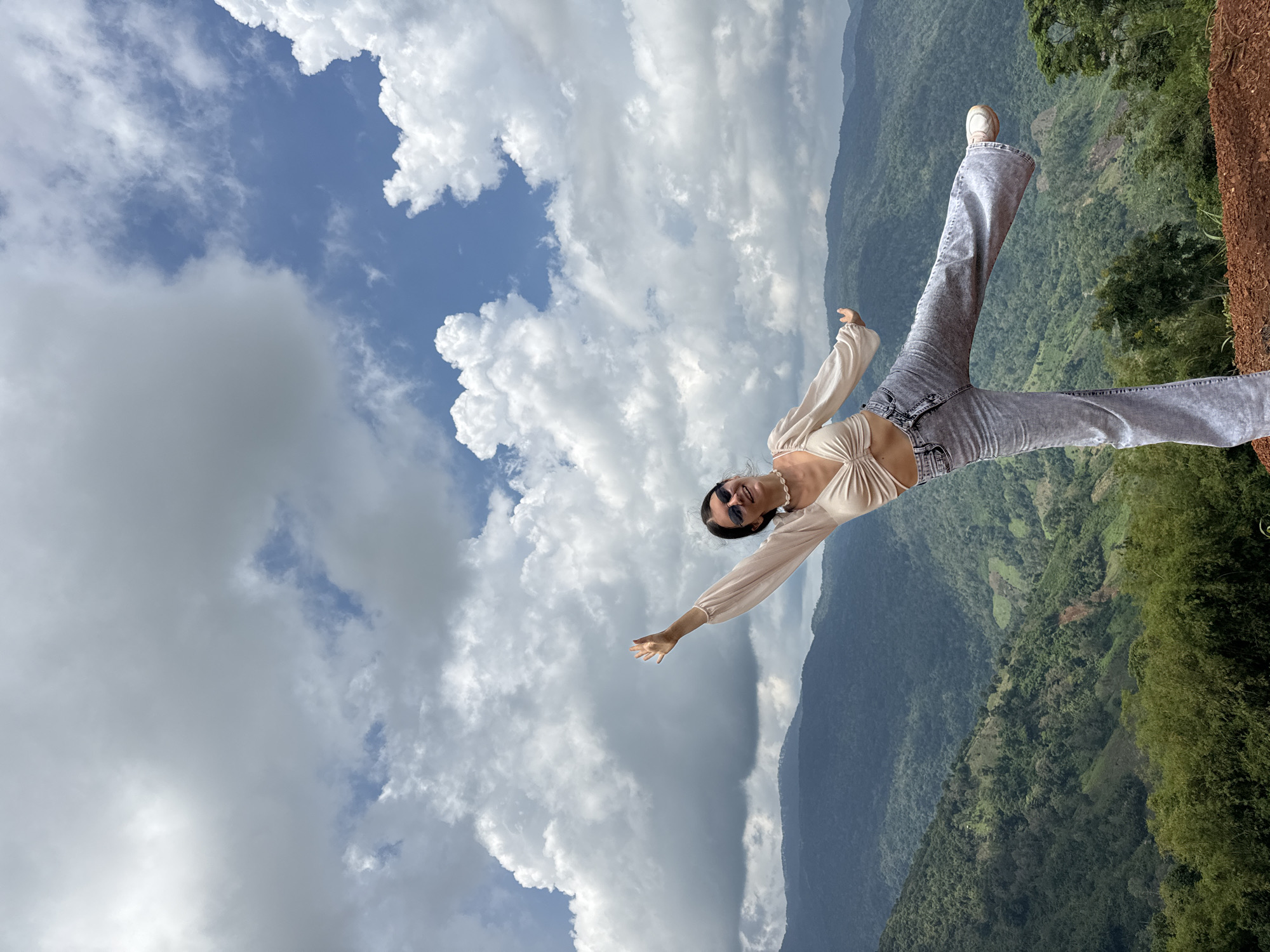 A young woman with dark hair, wearing a beige crop top, light gray flared jeans, and white sneakers, strikes a dynamic pose on a hill overlooking a valley. Her left leg is extended backward, and her right arm is raised in the air, with her left arm outstretched to the side. She wears sunglasses and a shell necklace. The background features rolling, tree-covered mountains under a cloudy blue sky. The foreground consists of reddish-brown earth and some green foliage. The location appears to be in the mountainous region of Nao Sẻ, Vietnam, judging by the distinct terraced landscape and lush greenery visible in the distance.
